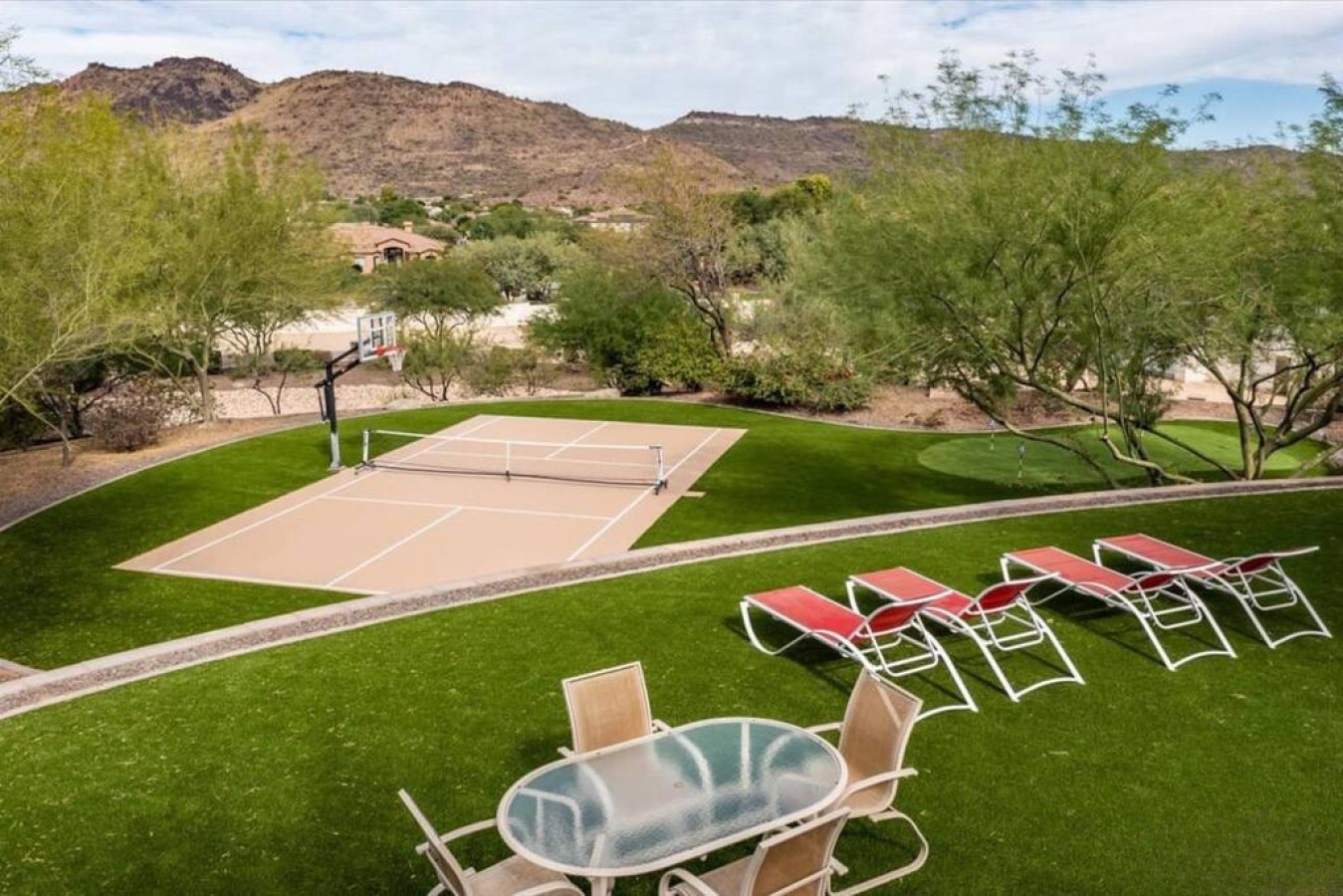 Private tennis court and lounge area set against desert mountain scenery with lush green lawn and mature trees framing the space.