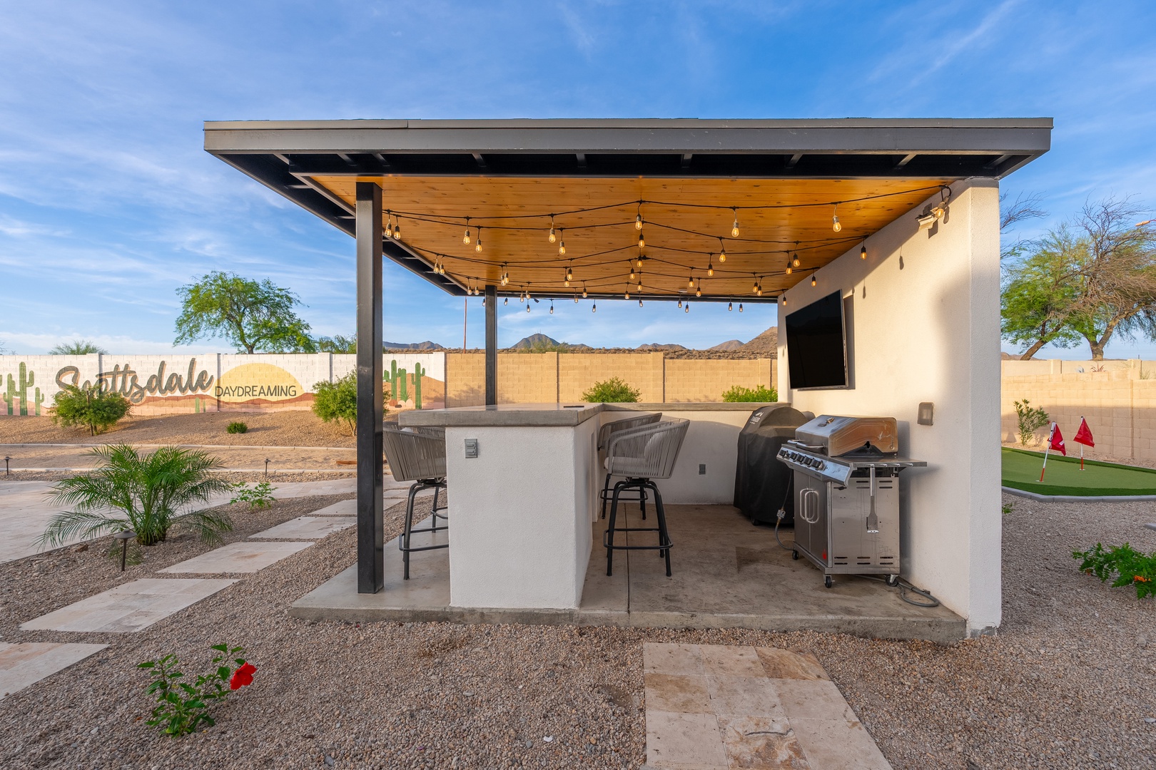 A modern covered patio features a barbecue station with bar seating, string lights overhead, and mountain views—perfect for evening gatherings under the stars.