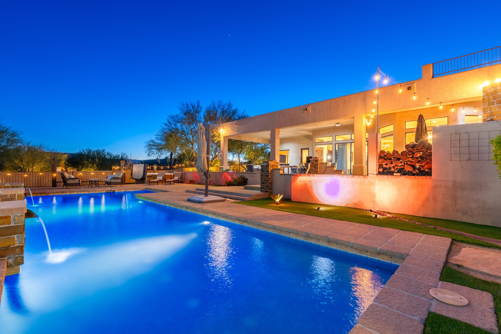 Stunning twilight view showcasing the property's illuminated modern architecture, expansive pool with ambient lighting, covered patio, and beautifully landscaped desert grounds.