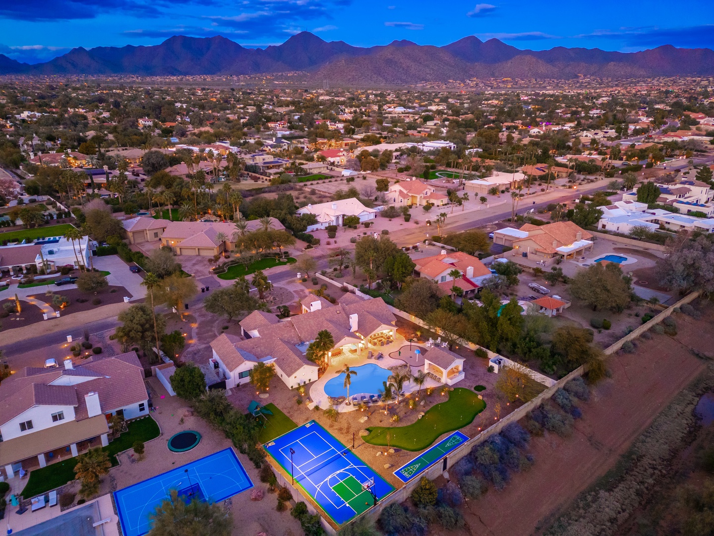 Aerial view of the property at dusk, showcasing the expansive desert setting with mountain ranges in the distance. Features private pool, tennis court, and sports facilities within a spacious residential neighborhood.