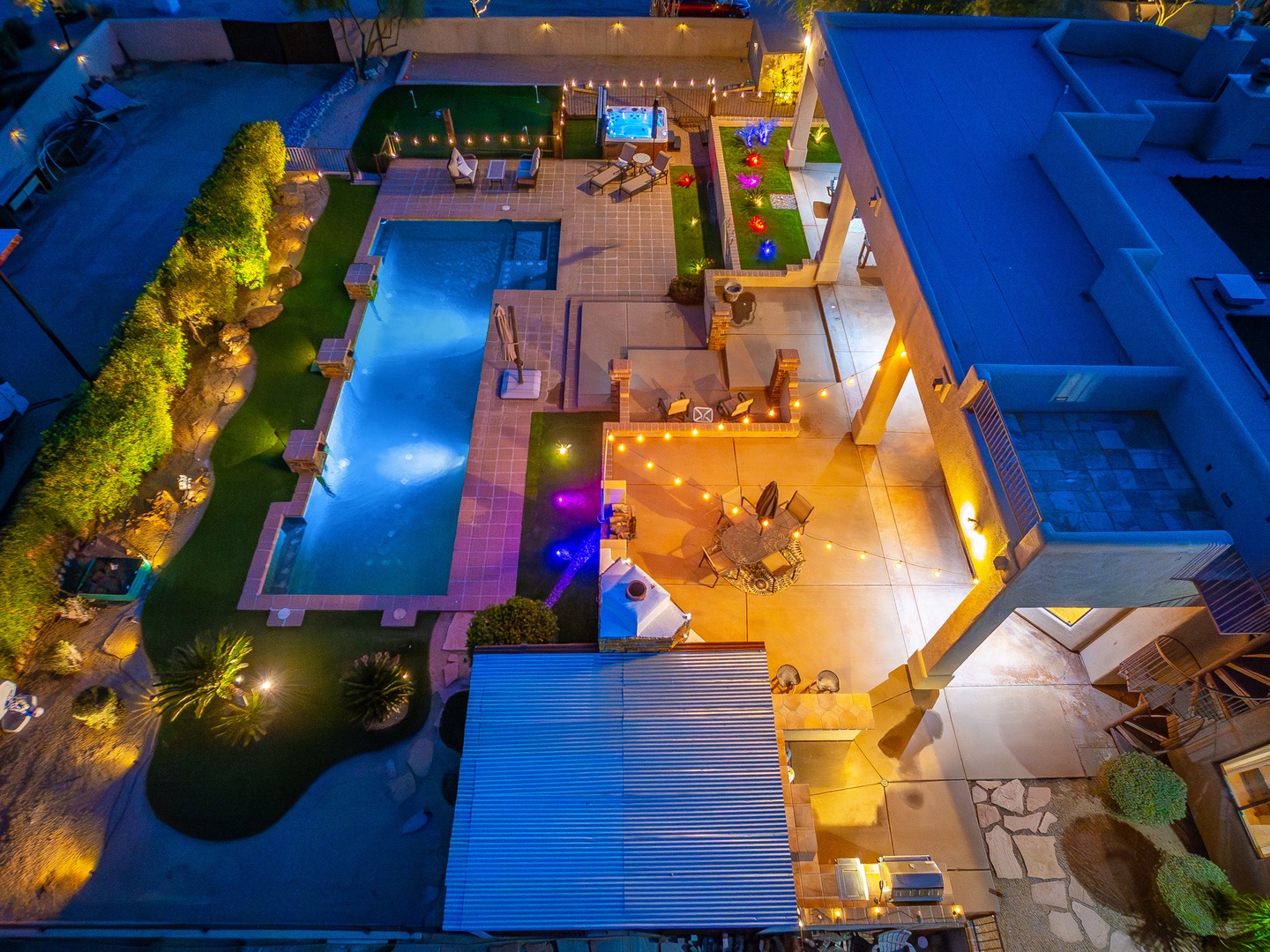 An aerial view captures a beautifully illuminated vacation rental at dusk, featuring a sparkling pool surrounded by lush landscaping, multiple outdoor entertaining spaces with string lights, and lounge areas perfect for evening gatherings.