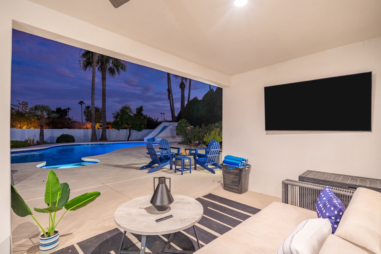 Your open-air living space flows seamlessly to a sparkling pool surrounded by palms, where blue chairs invite sunset lounging under the desert sky.