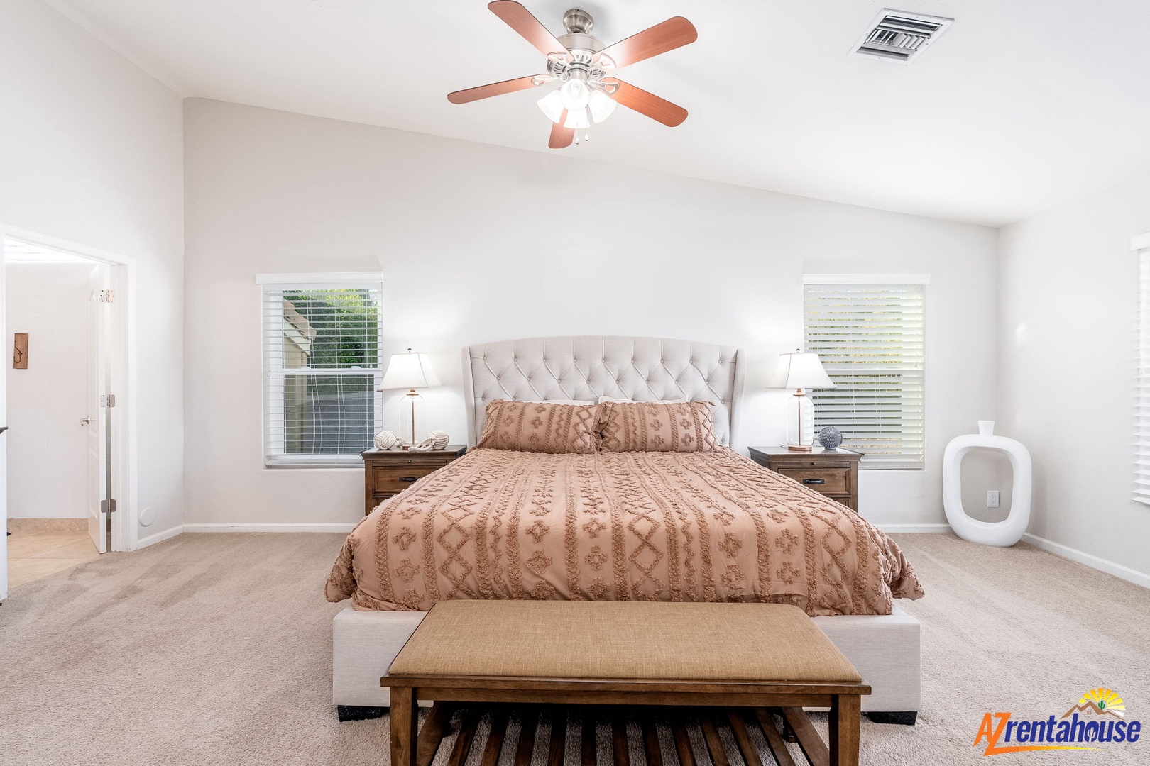 Your serene bedroom awaits with a plush upholstered bed, soft lighting, ceiling fan for comfort, and natural light from dual windows creating a peaceful retreat.
