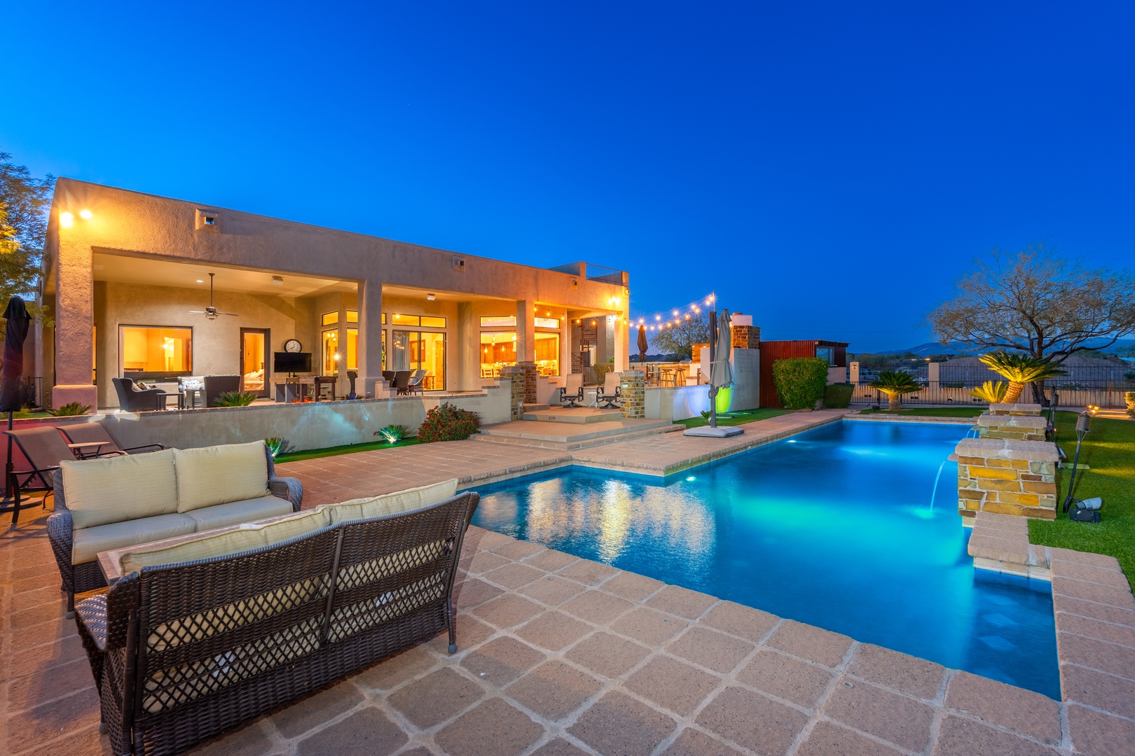 A stunning Southwestern-style property featuring an illuminated resort-style pool with spa, expansive covered patio, mountain views, and elegant desert landscaping, photographed at dusk.