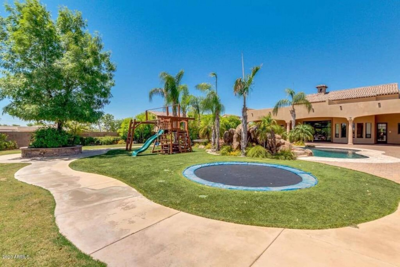 Expansive backyard paradise featuring playground equipment, in-ground trampoline, swimming pool, and palm-tree landscaping under clear Arizona skies.