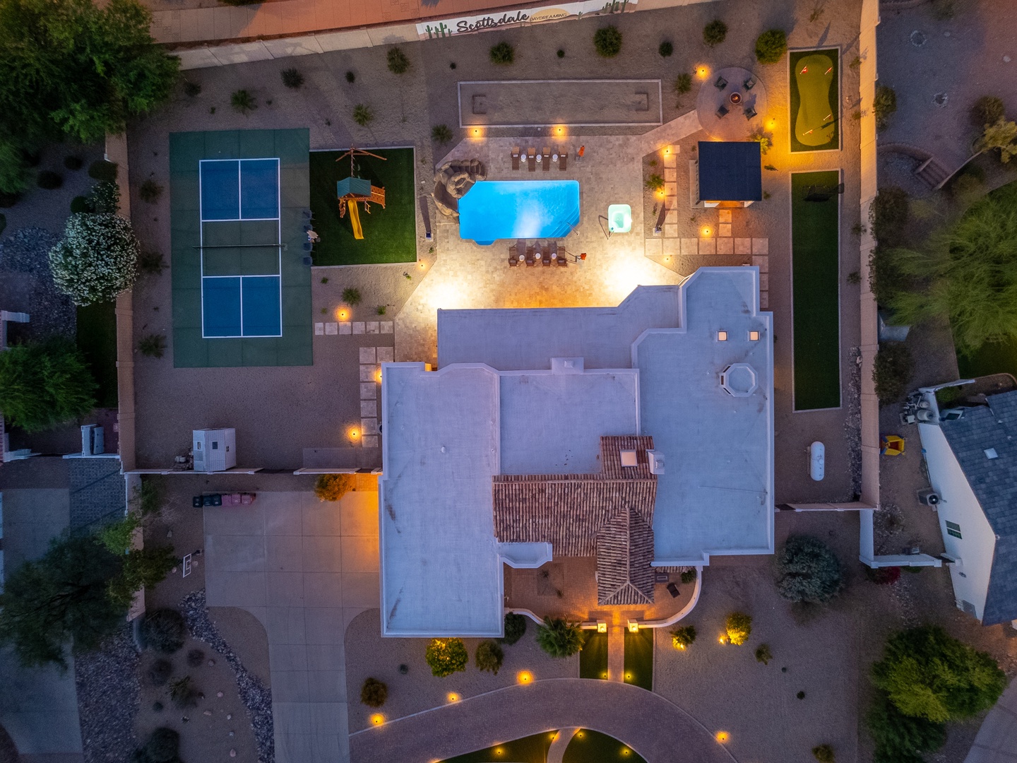 Aerial view of the property at dusk showcasing a spacious layout with a central swimming pool, tennis court, landscaped grounds, and multiple recreational areas including a putting green and outdoor seating spaces.