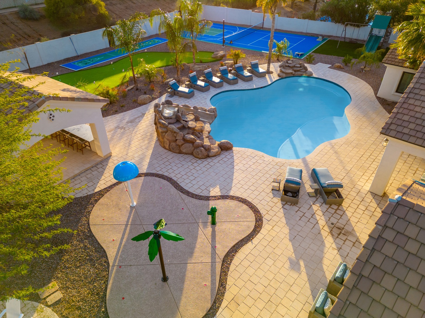 An aerial view showcasing the property's expansive outdoor recreation area with a curved pool, sports court, and tropical landscaping framed by palm trees and sun loungers.