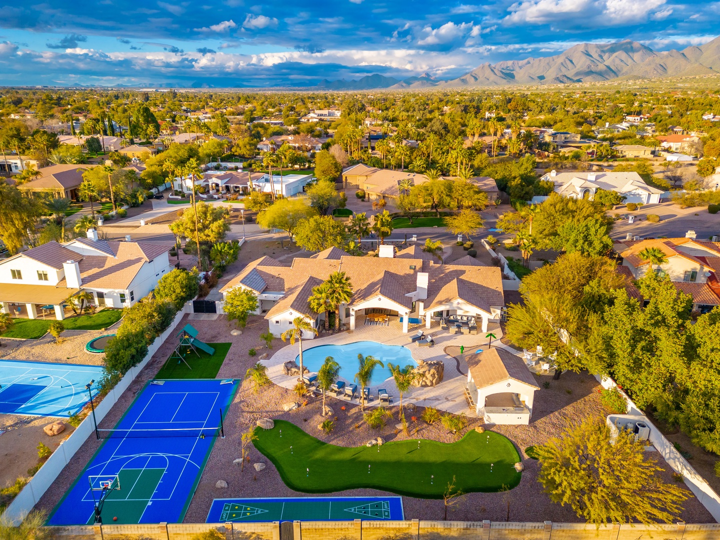 Aerial view showcasing a stunning desert retreat in a residential neighborhood with mountain vistas, featuring a resort-style pool, mini golf, sport court, and generous outdoor entertainment spaces.