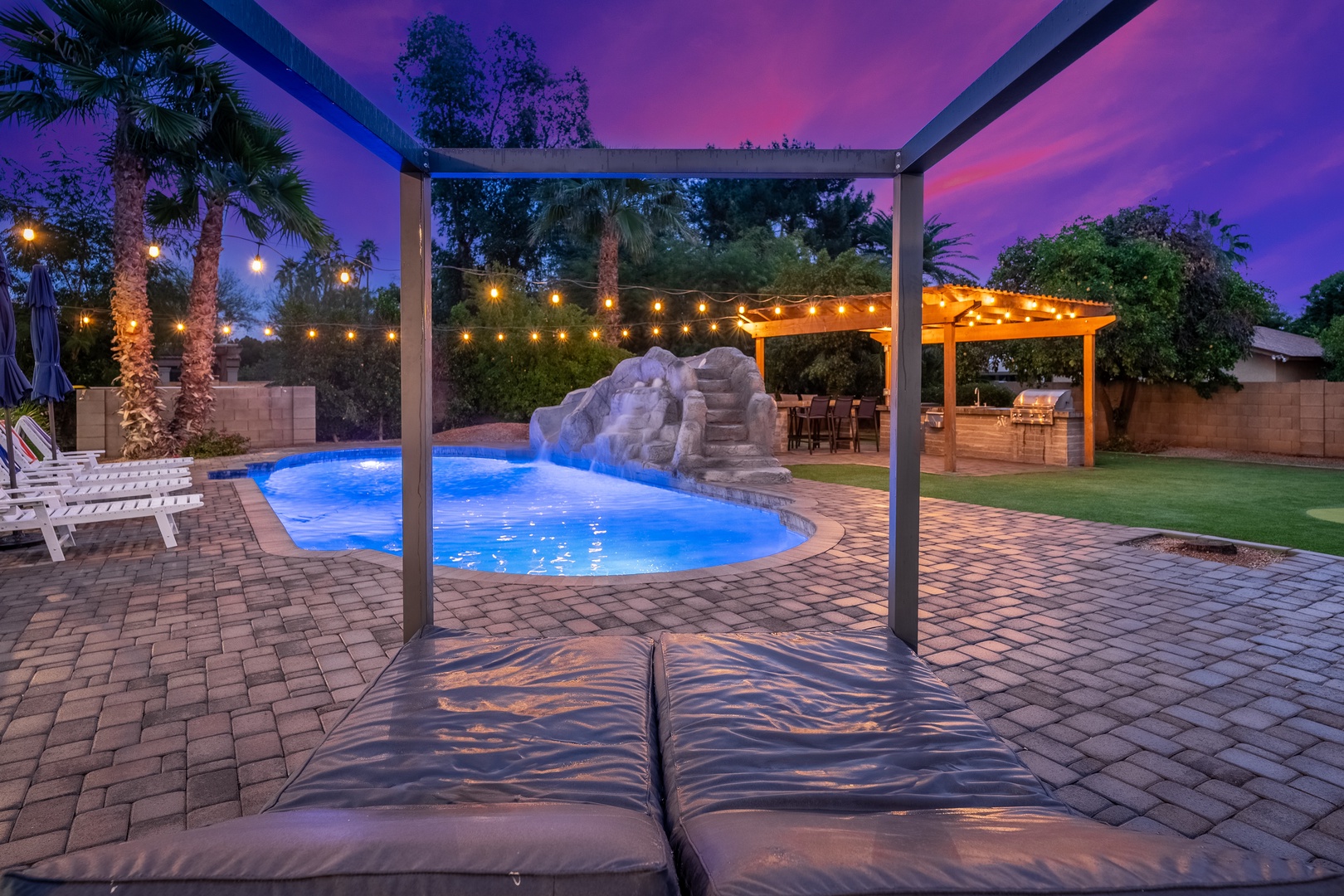 A vibrant desert evening painted in purple and pink hues frames the inviting backyard retreat featuring a sparkling pool with waterfall rocks, string-lit pergola dining, and lush palm trees.