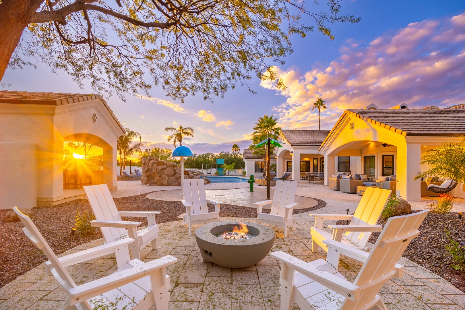 The property features an expansive outdoor living area with a fire pit, multiple covered patios, pool amenities, and dramatic mountain vistas, all set beneath desert skies at sunset.