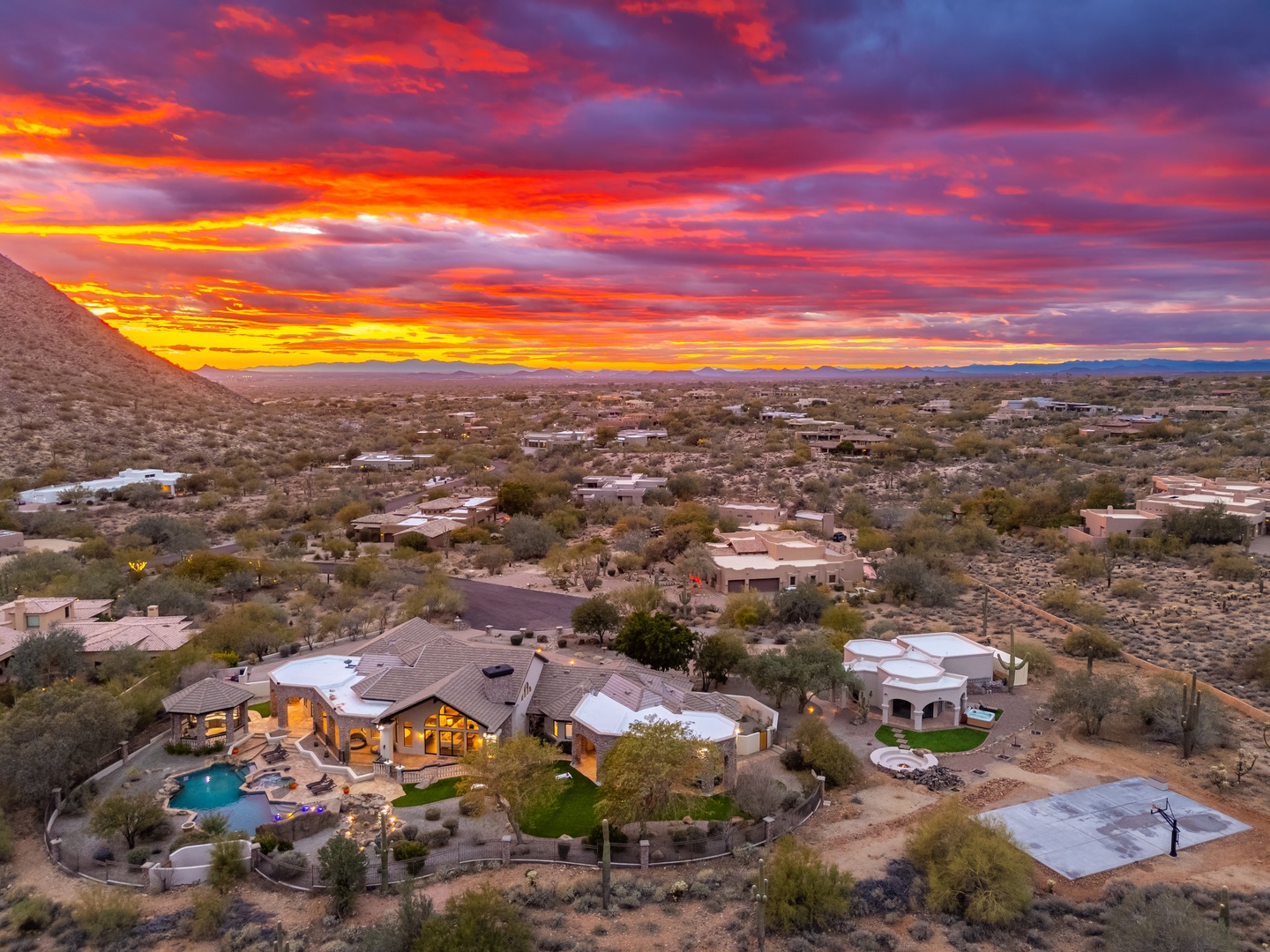 Aerial view captures the property nestled in the desert landscape, with dramatic sunset skies painting the horizon in brilliant oranges, reds, and purples over the surrounding community.