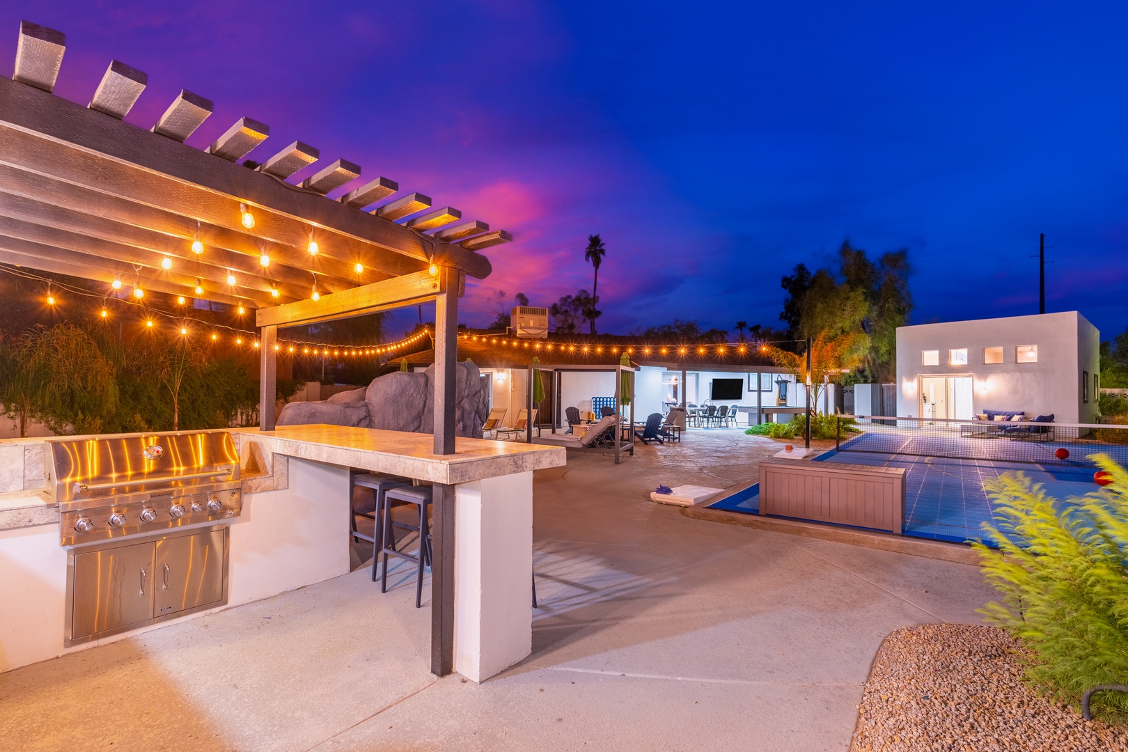 A stunning modern property with outdoor pool, BBQ facilities, and warm string lighting set against a vibrant desert sunset sky.