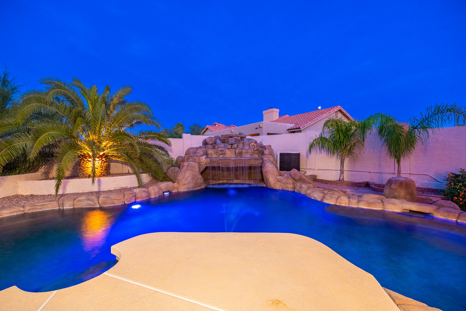 Stunning pool illuminated at twilight, featuring a cascading rock waterfall, tropical palms, and ambient lighting that creates a serene evening atmosphere in the backyard.