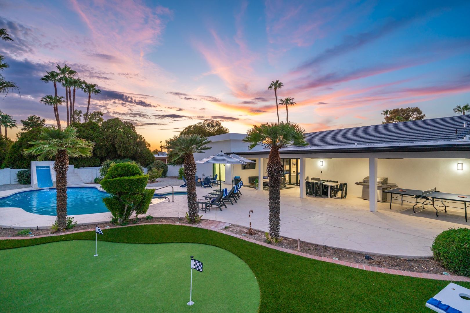 A stunning private retreat showcasing a sparkling pool, putting green, covered patio with dining and ping-pong, and tropical landscaping under a breathtaking desert sunset sky.