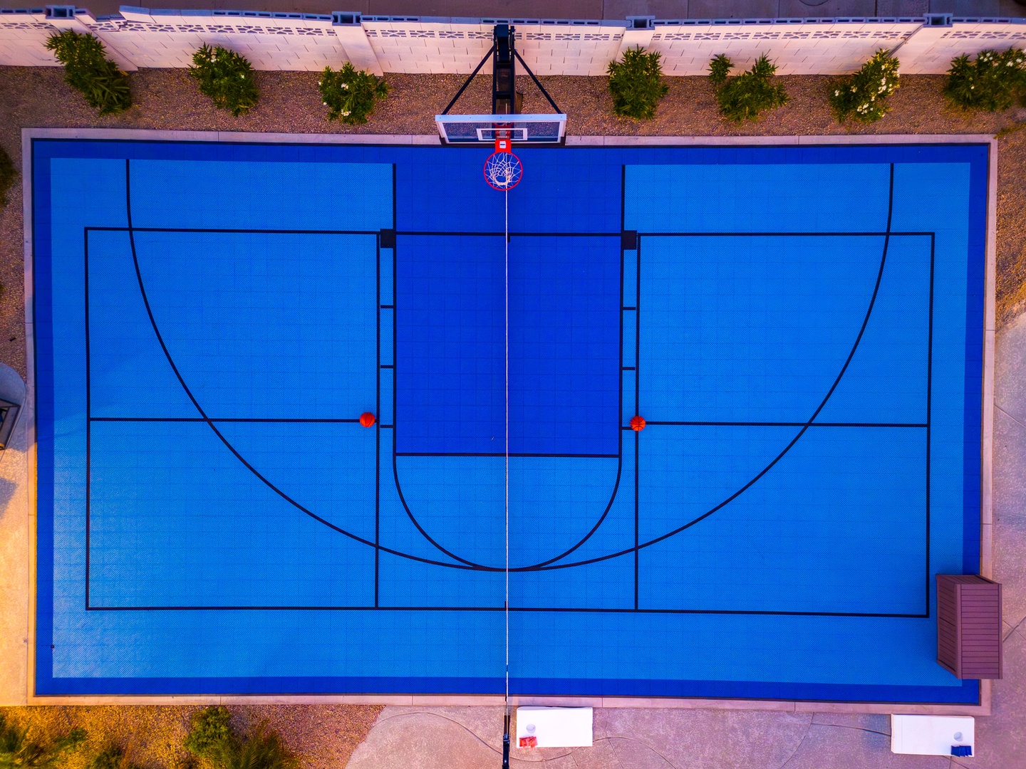 An aerial view of a vibrant blue full-size basketball court with marked lines, a center hoop, and landscaped surroundings at the property.