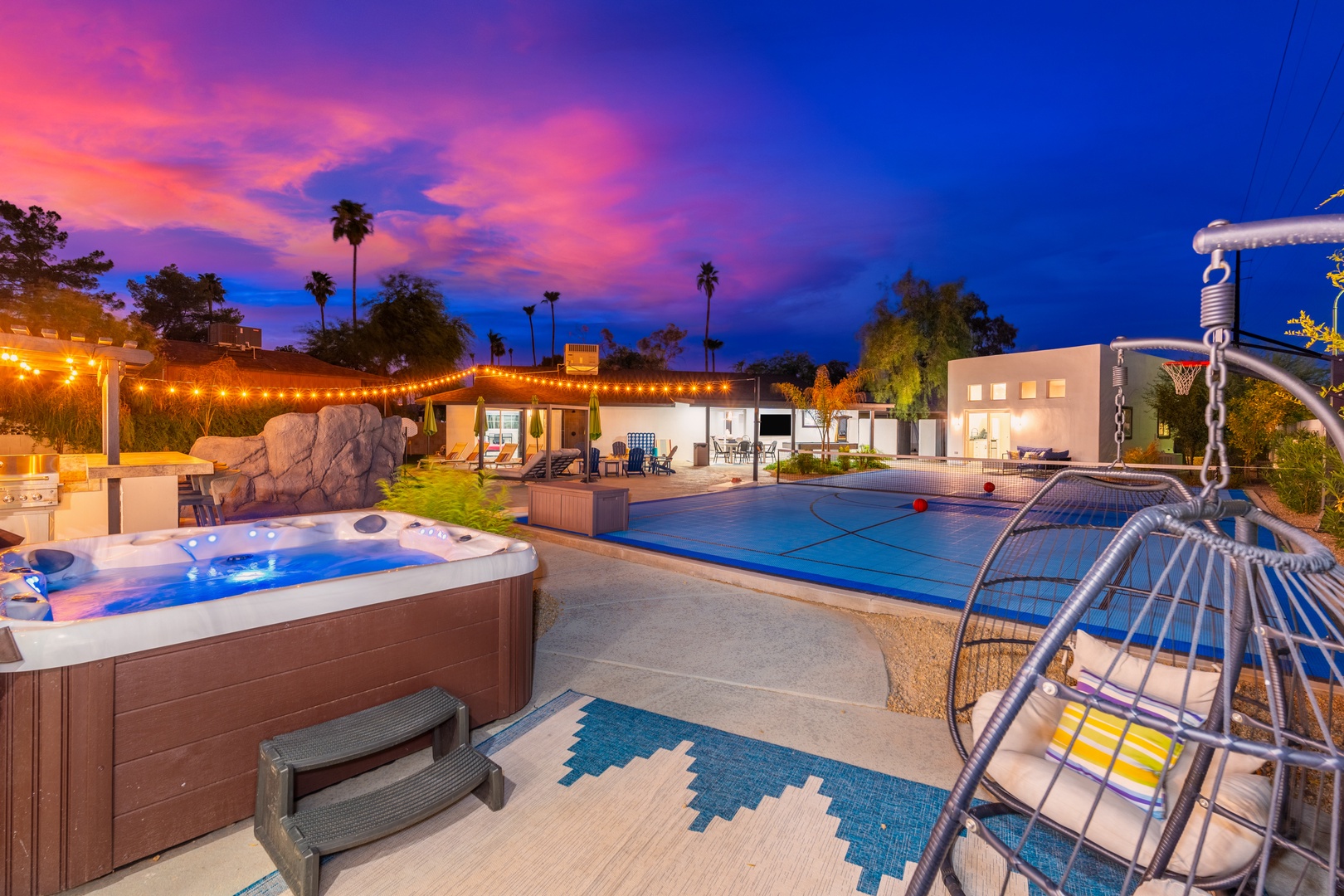 A stunning desert retreat featuring a hot tub, pool, and outdoor lounge area, all framed by palm trees and a dramatic pink and purple sunset sky.