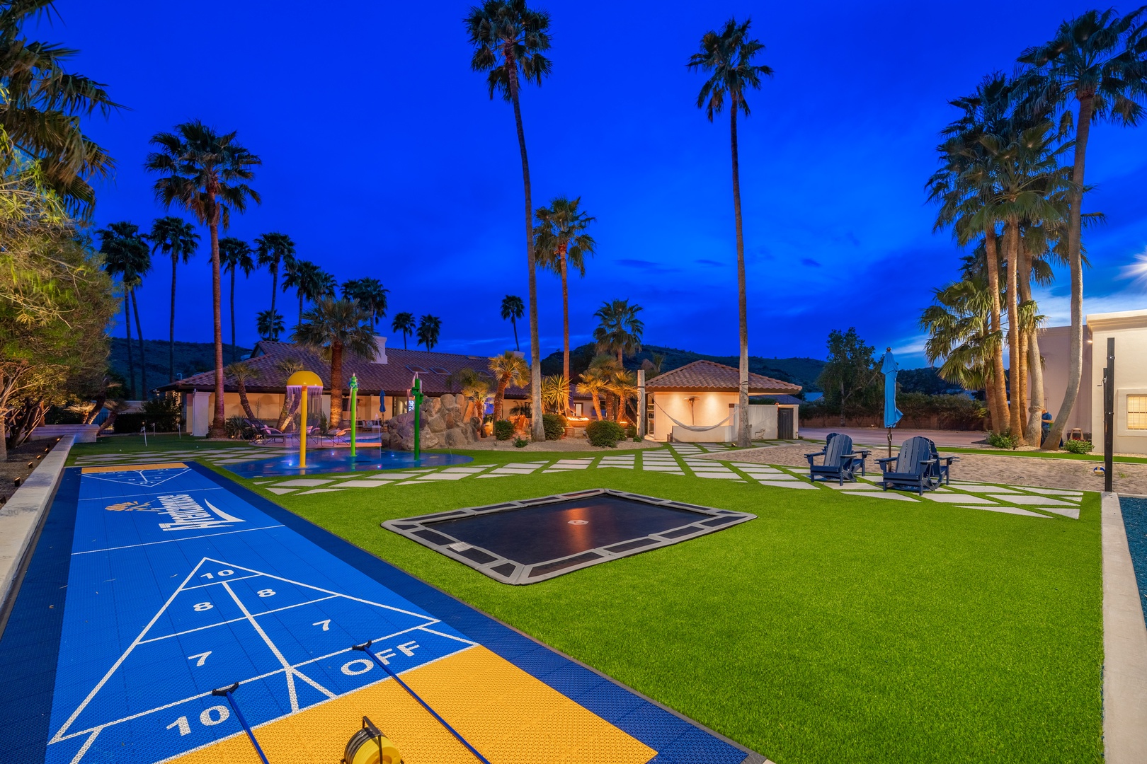 A sprawling desert resort property features multiple guest bungalows and recreational areas beneath towering palm trees at dusk, with mountains visible in the distance.
