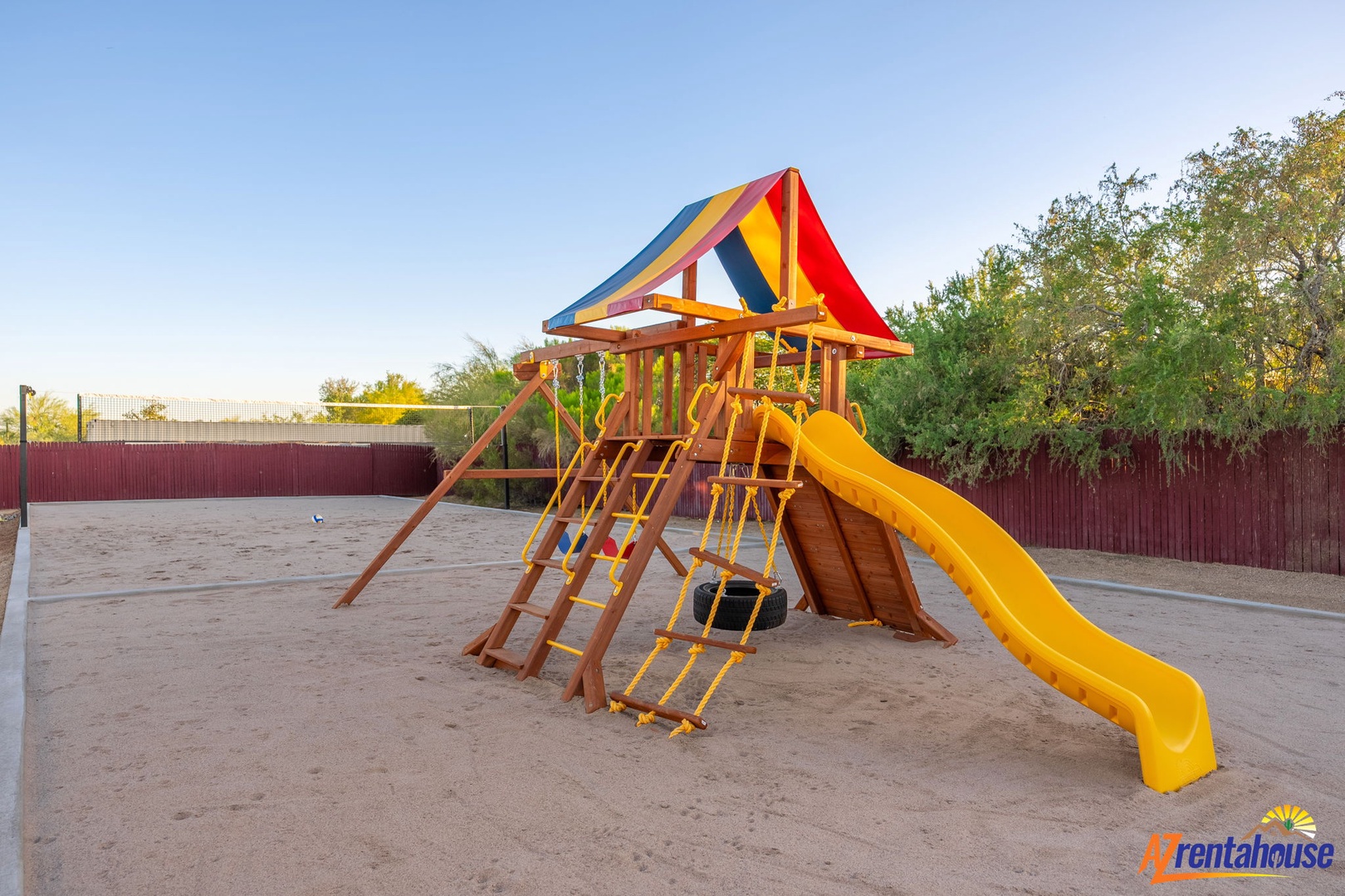 A colorful playground with a bright yellow slide and climbing features awaits young adventurers in a safe, sandy play area surrounded by trees and open space.