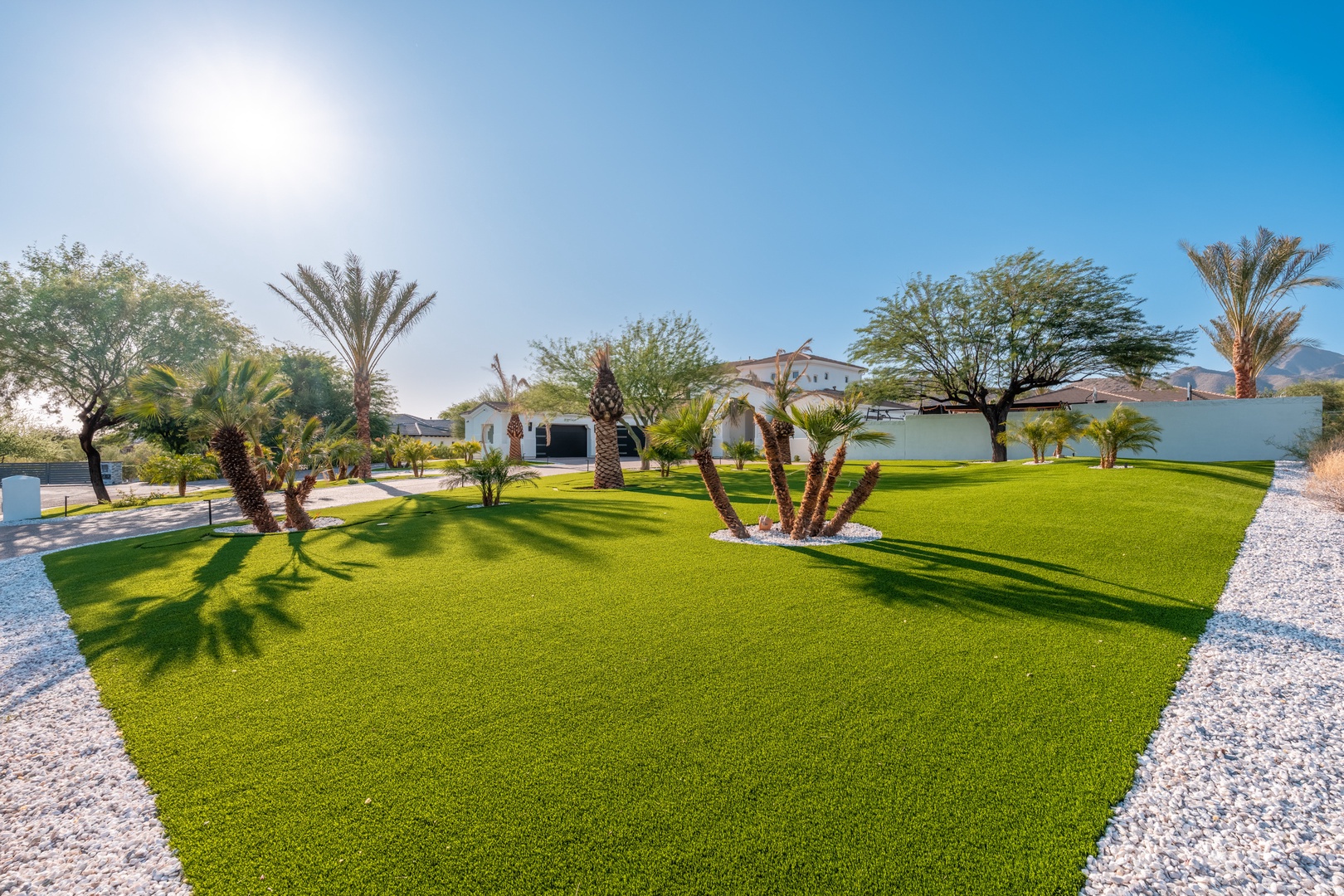 A meticulously landscaped property with lush green lawns, mature palm trees, and manicured gardens set against mountain views. The exterior showcases modern design with gravel pathways and extensive outdoor grounds.