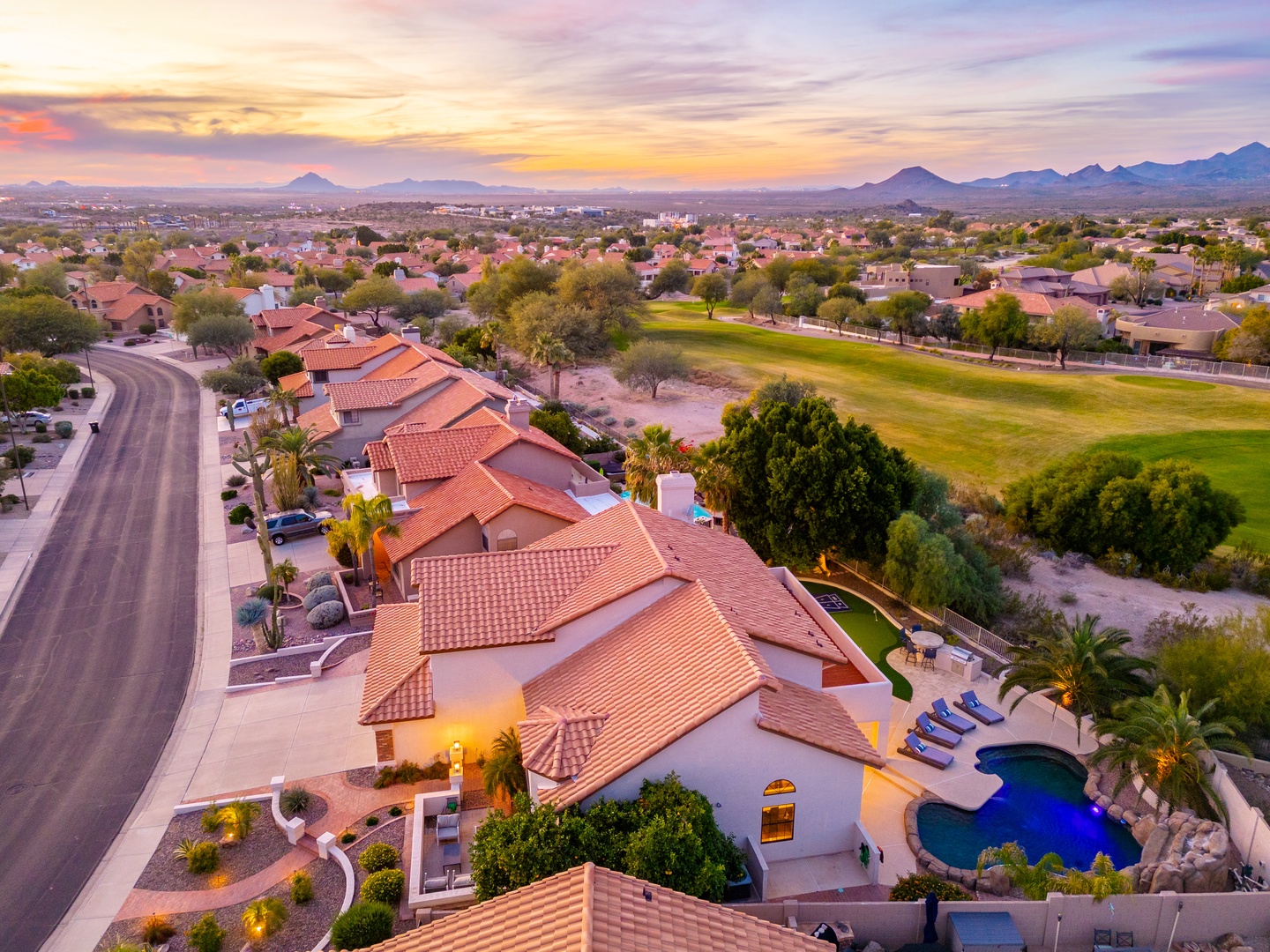 Aerial view of the property nestled in a scenic desert community, with a private pool, lush golf course greens nearby, and mountain ranges framing the horizon at sunset.