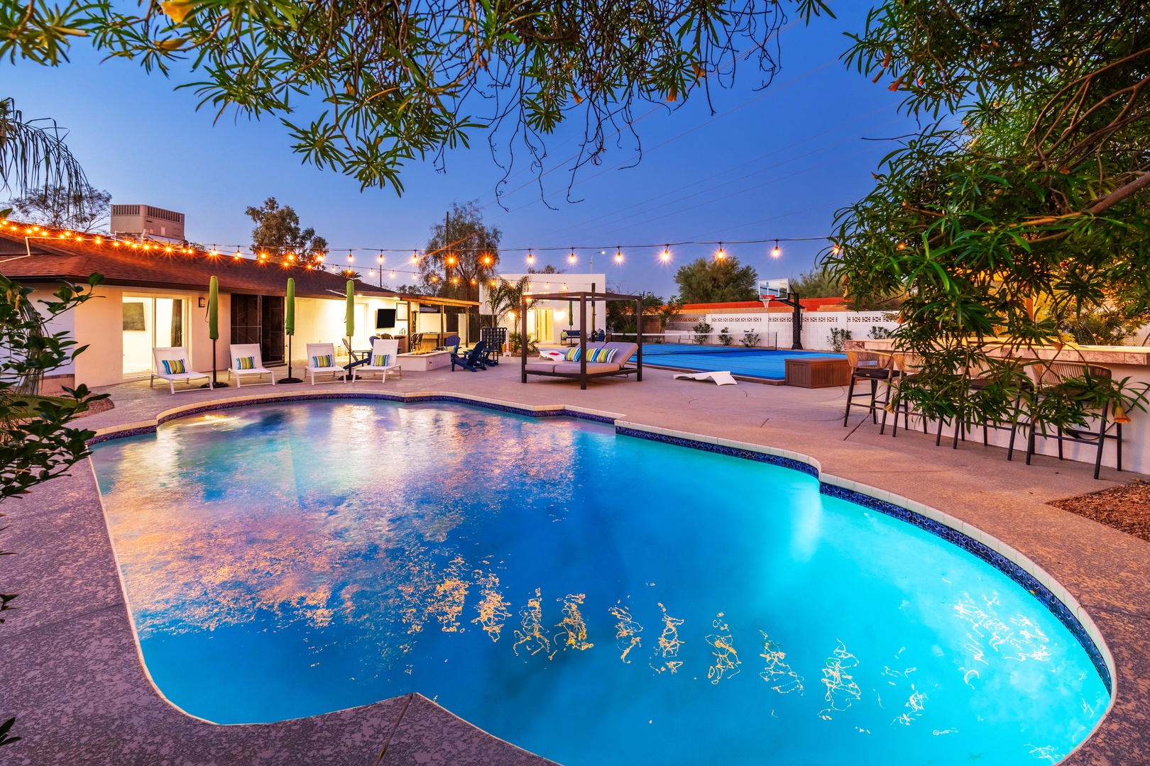 Tranquil evening retreat featuring two sparkling pools surrounded by lush landscaping, ambient string lighting, and multiple relaxation areas perfect for unwinding under the desert sky.