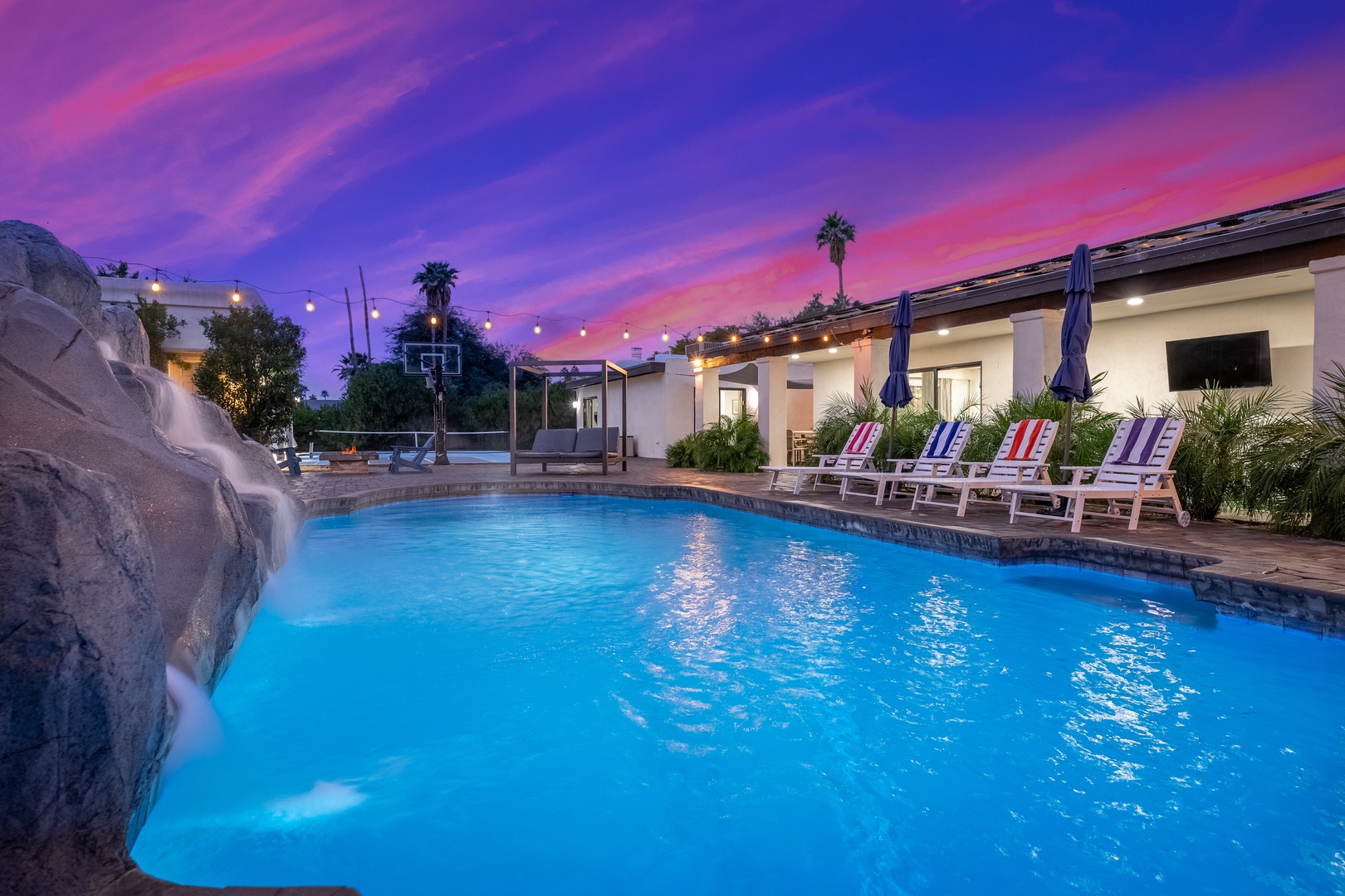 Stunning sunset paints the sky as you unwind poolside, with lounge chairs ready, waterfall feature, basketball court nearby, and string lights overhead setting a magical ambiance.