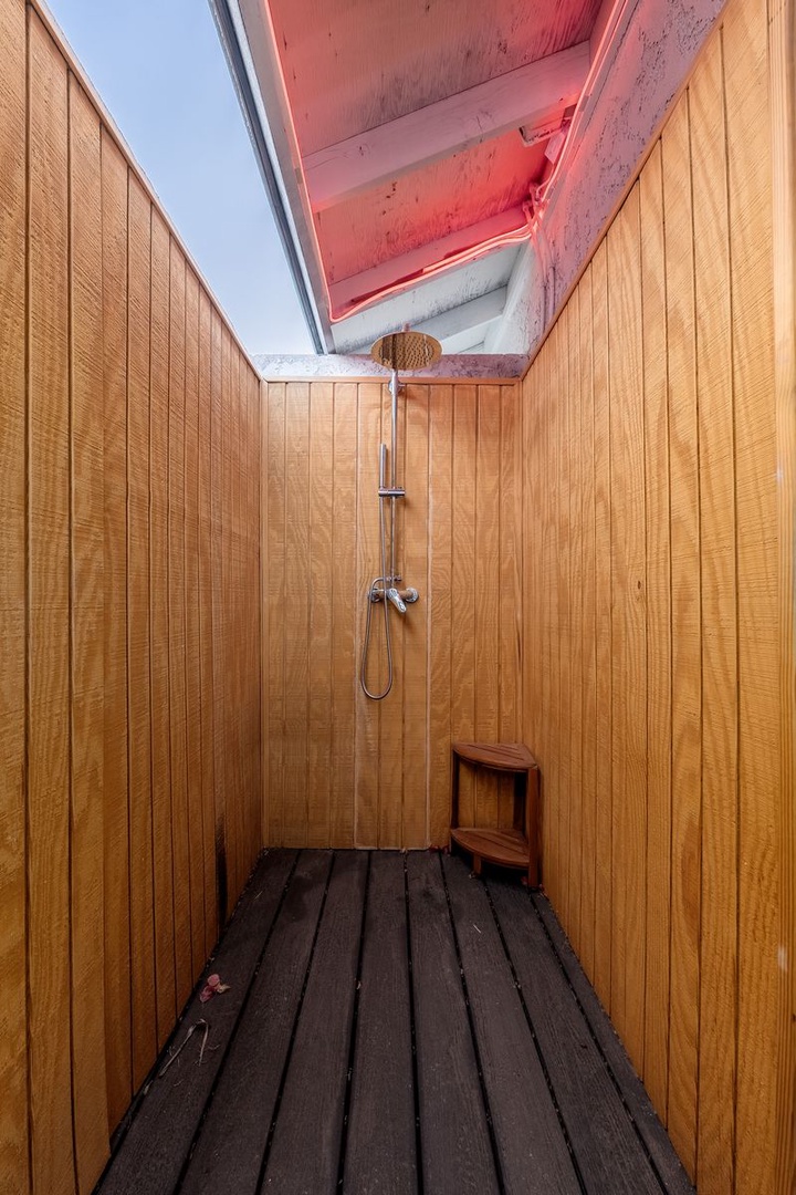 An open-air shower featuring warm wood paneling, wooden flooring, and a distinctive pink-accented skylight overhead for a unique outdoor bathing experience.