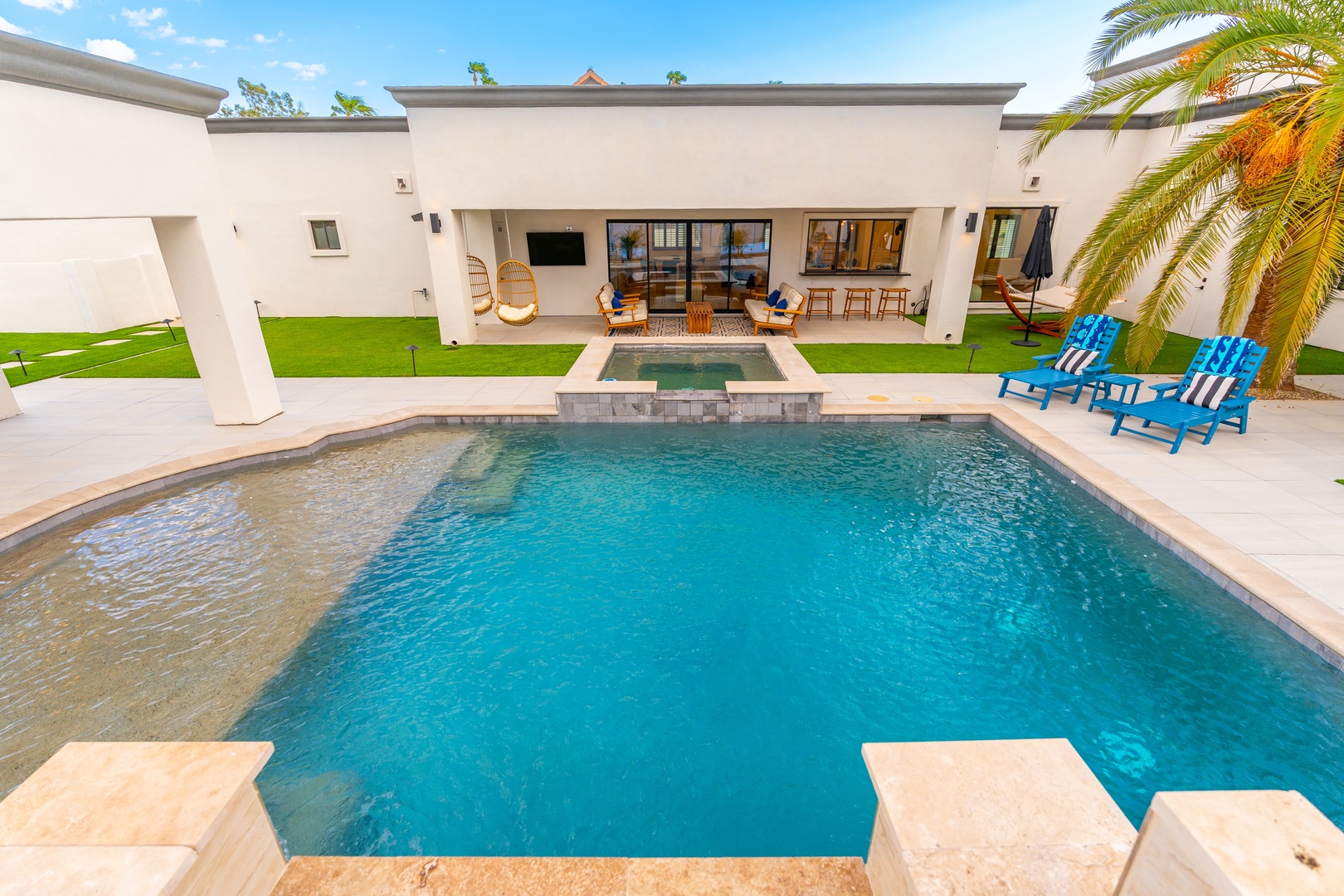 Modern desert retreat with sparkling pool, spa, vibrant lounge chairs, and covered patio beneath swaying palms—your perfect sunny escape awaits.