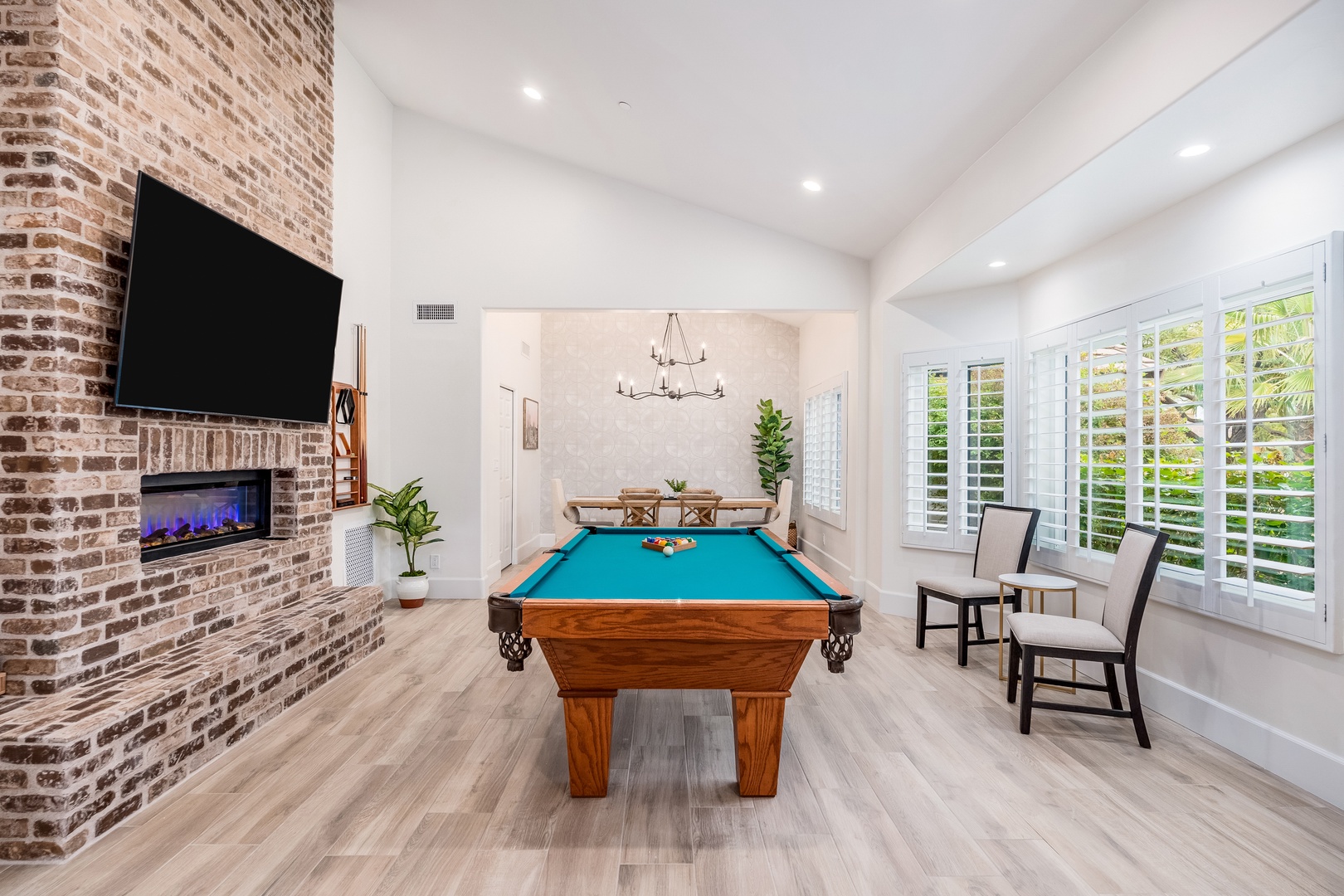 Gather for friendly competition in your game room, where billiards await beneath soaring ceilings, framed by a rustic brick fireplace and sunlight streaming through plantation shutters.