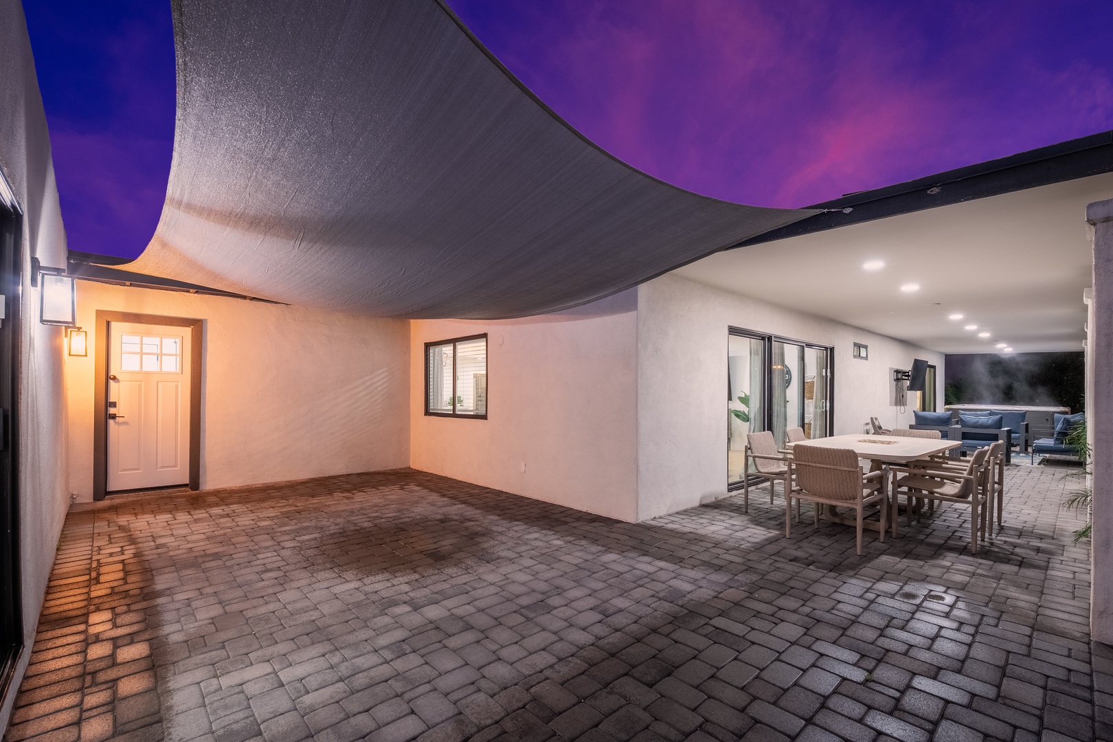 This covered patio area features vibrant purple shade sails above brick pavers, creating an inviting entrance space with ambient lighting and a separate outdoor dining area visible in the background.
