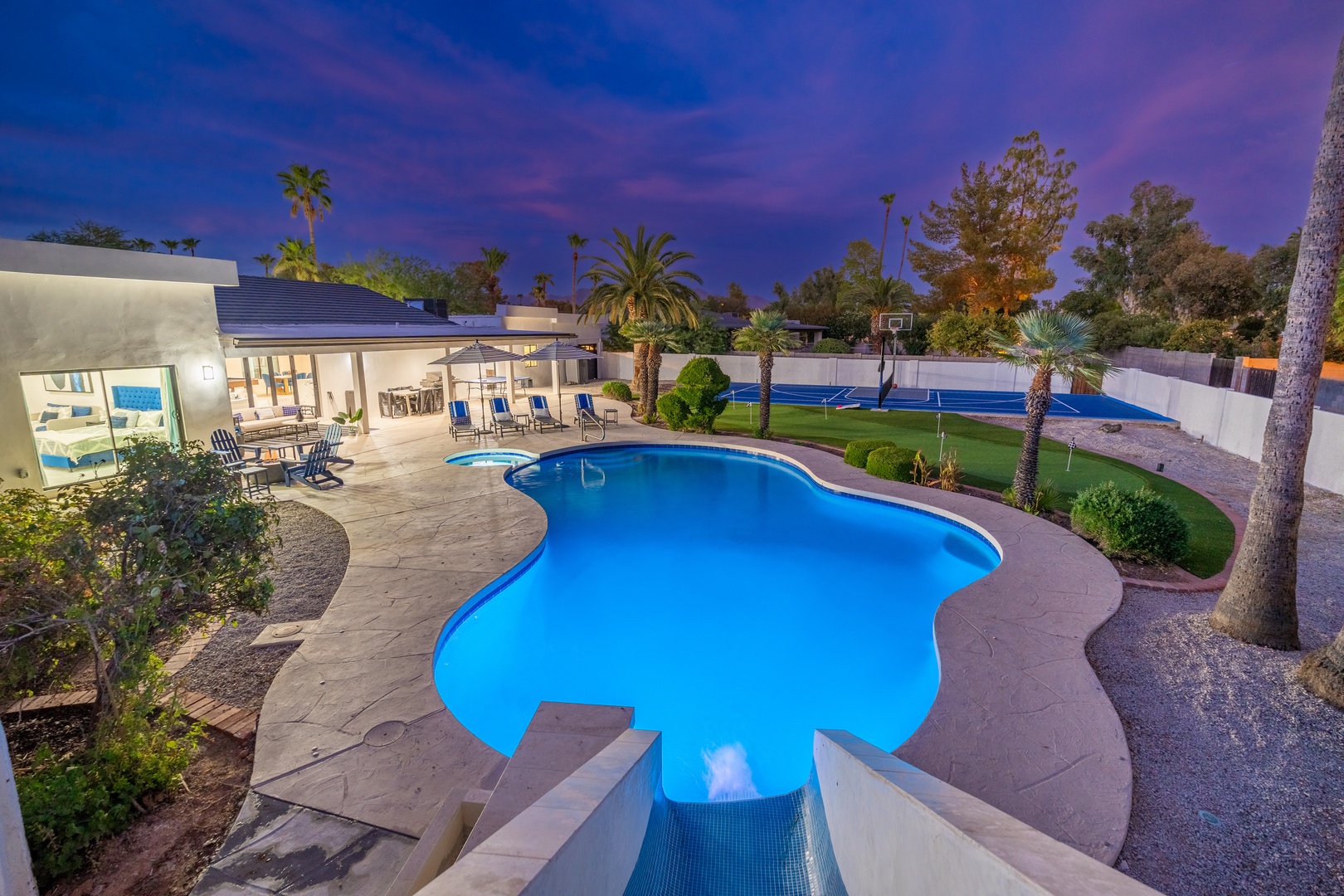 The illuminated pool and spacious backyard framed by palm trees create an inviting oasis for evening relaxation under a stunning twilight sky.