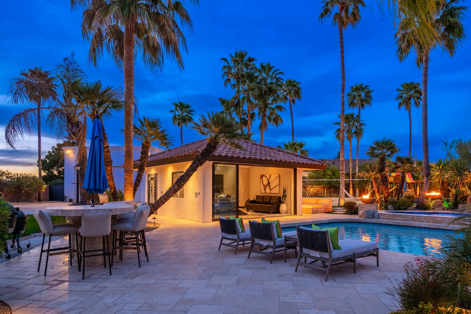 Modern desert oasis at dusk, featuring a sparkling pool, palm-lined grounds, outdoor dining and lounge areas beneath a dramatic twilight sky.