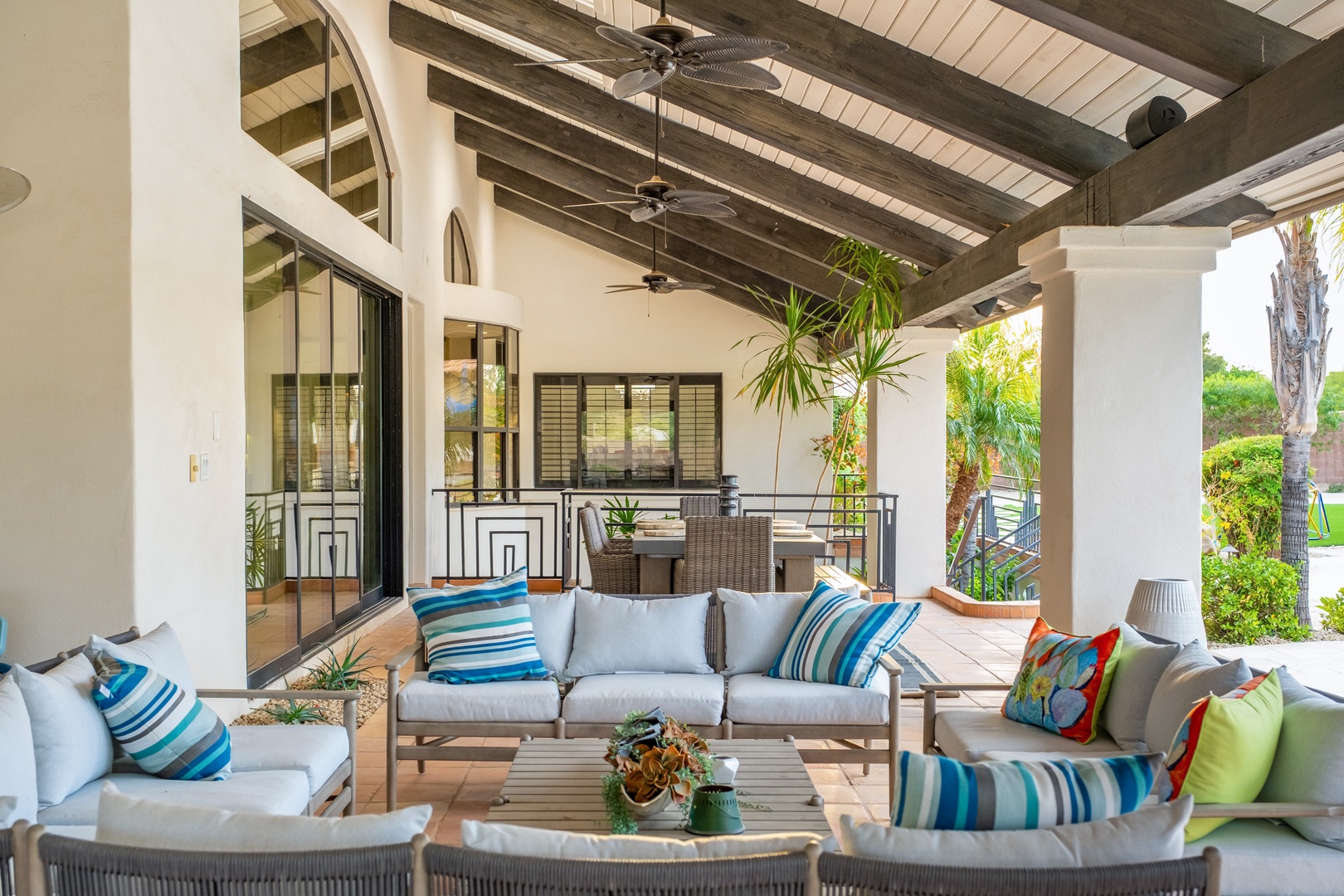Settle into plush seating beneath the ceiling fans and dark wood beams of your covered patio, surrounded by tropical greenery and garden views—perfect for relaxing or hosting.