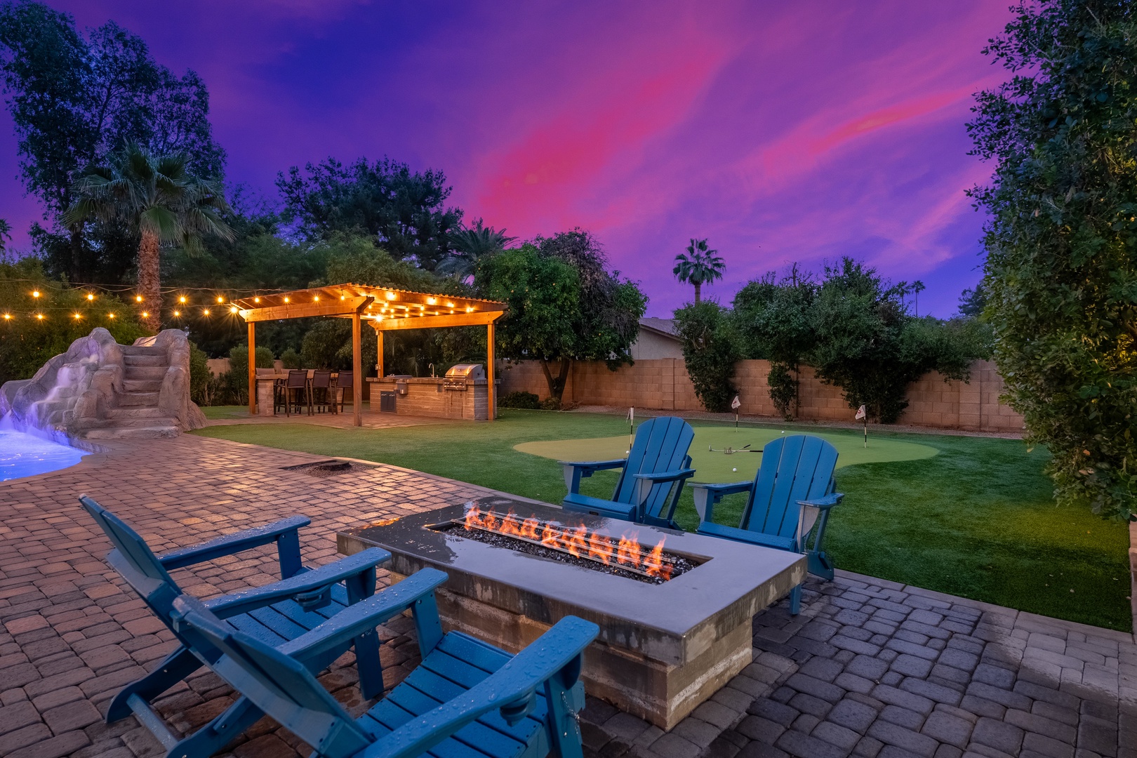 A stunning backyard retreat featuring a custom firepit surrounded by Adirondack chairs, manicured putting green, illuminated pergola with dining area, and tropical landscaping beneath a vibrant twilight sky.