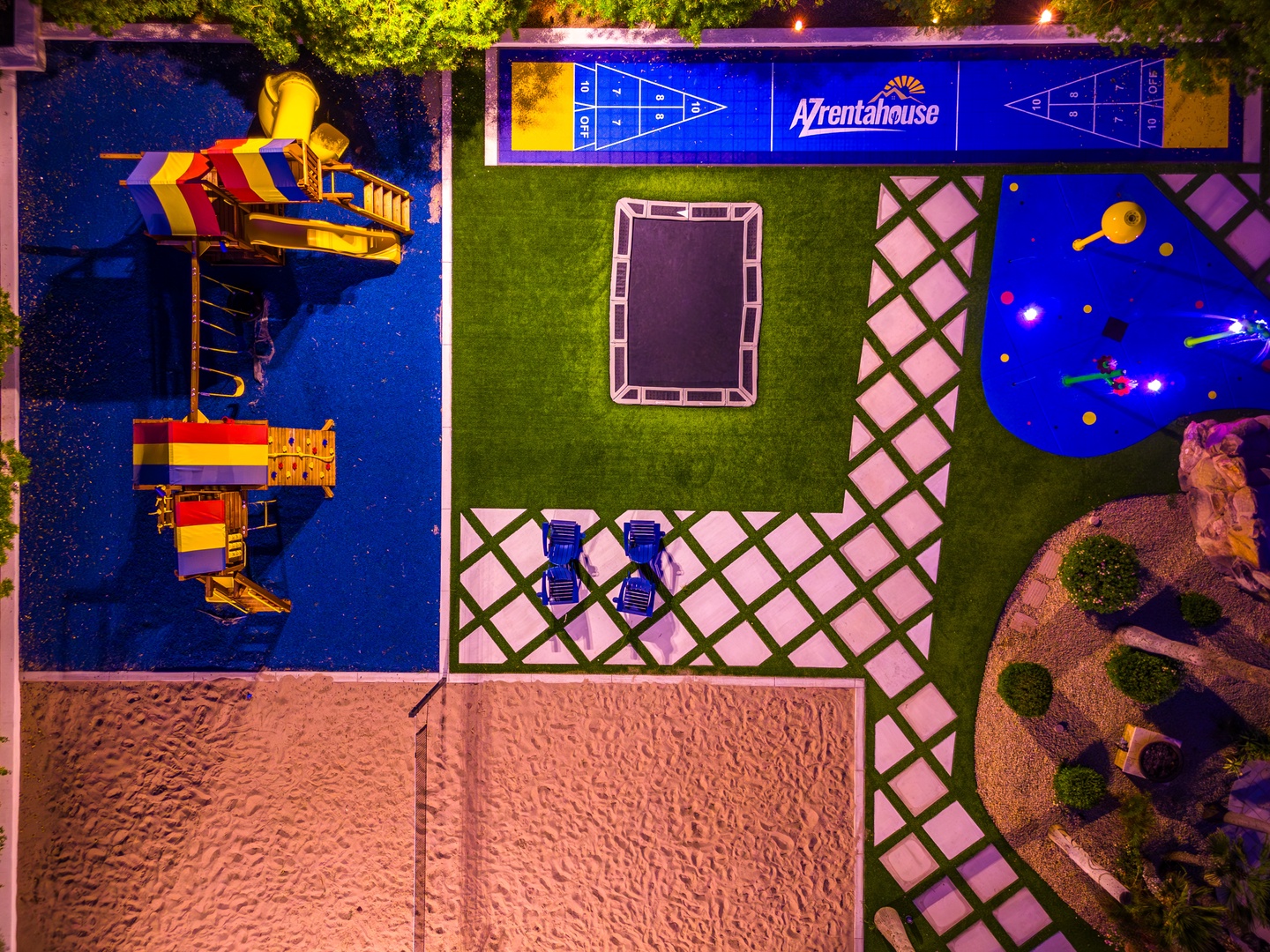 Aerial view of AZrentahouse property showing vibrant pools, playground equipment, shuffleboard court, private trampoline, sun loungers, sandy beach area, and beautifully landscaped grounds illuminated at night.