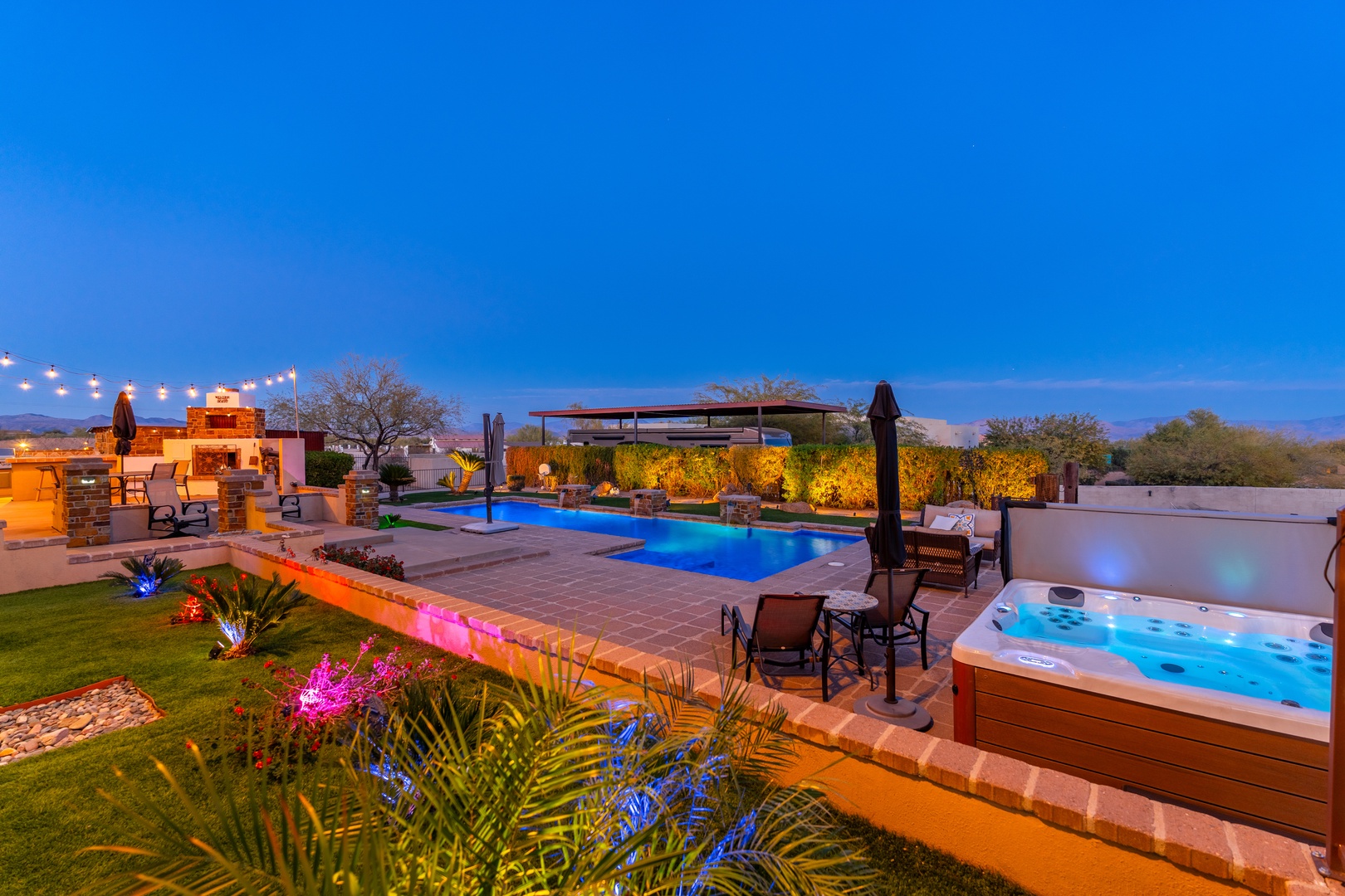 An outdoor oasis awaits at twilight, featuring a sparkling pool, relaxing hot tub, and illuminated lounge areas surrounded by lush landscaping and ambient lighting.