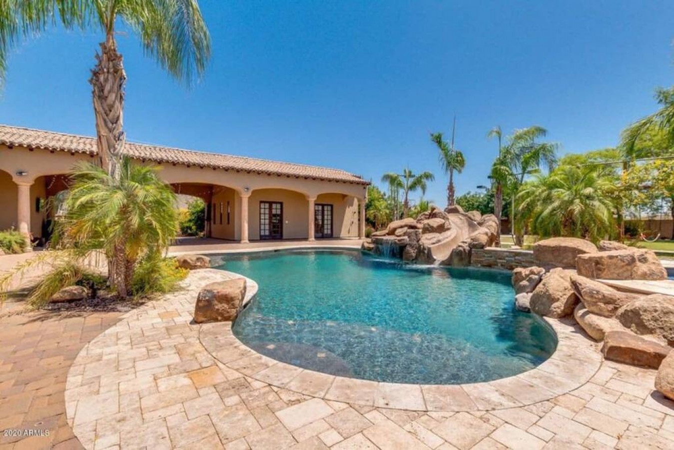 A stunning Mediterranean-style villa with a crystal-clear pool featuring a stone waterfall, surrounded by lush palm trees and pristine landscaping under bright blue skies.