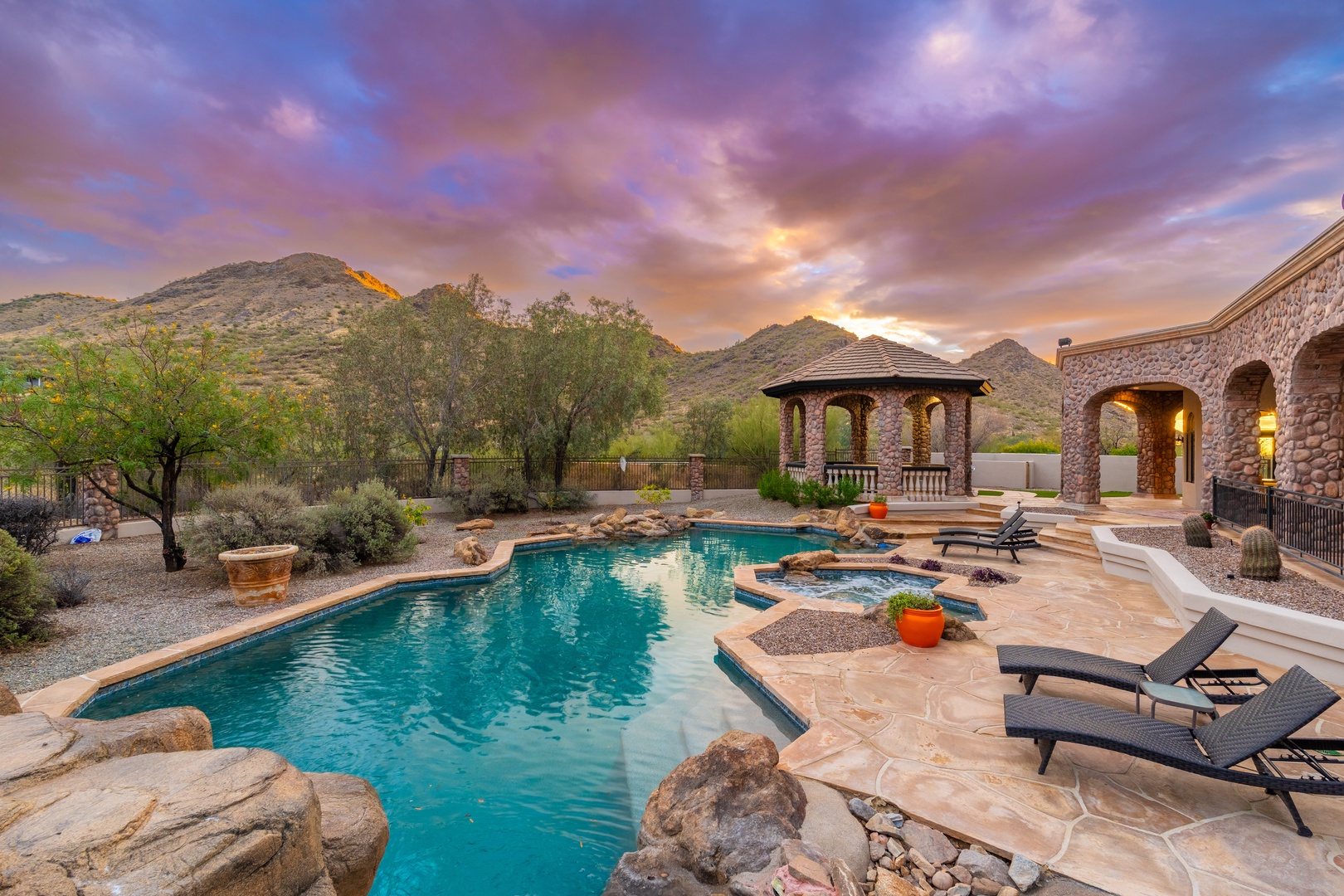 The property offers a private outdoor pool and spa area surrounded by desert landscaping, with a covered gazebo and mountain backdrop creating a serene setting during sunset hours.