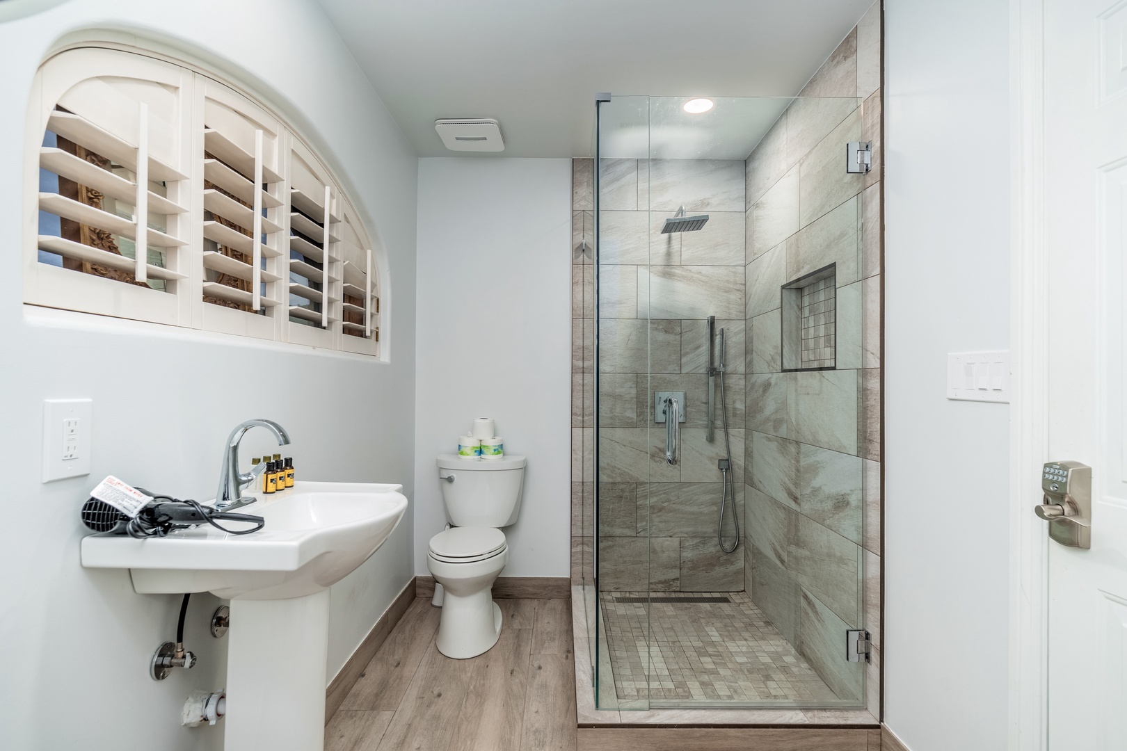 Your private bathroom features a spacious walk-in shower with sleek glass doors, modern fixtures, and soft natural light filtering through elegant shutters above the sink.