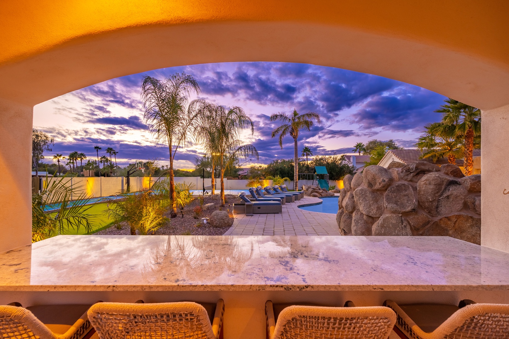 Luxurious desert retreat framed by archways, with stunning views of tropical palms, inviting pool areas, and dramatic skies at dusk. A perfectly landscaped outdoor oasis awaits.