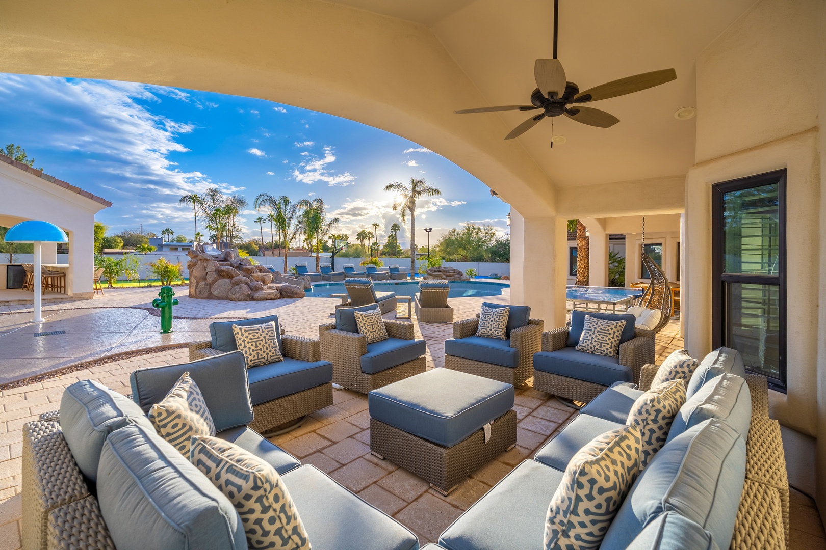 A spacious covered patio featuring plush seating and decorative pillows frames views of the sparkling pool, rock waterfall feature, and palm-dotted landscape—your perfect setting for alfresco lounging.