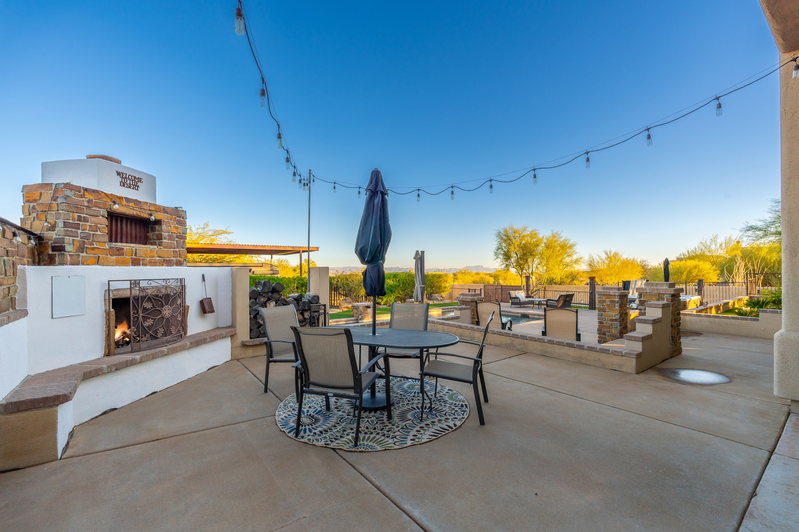 Spacious outdoor patio features an elegant stone fireplace, comfortable seating, and panoramic desert views under string lights—perfect for evening gatherings as the golden hour casts warm tones across the landscape.