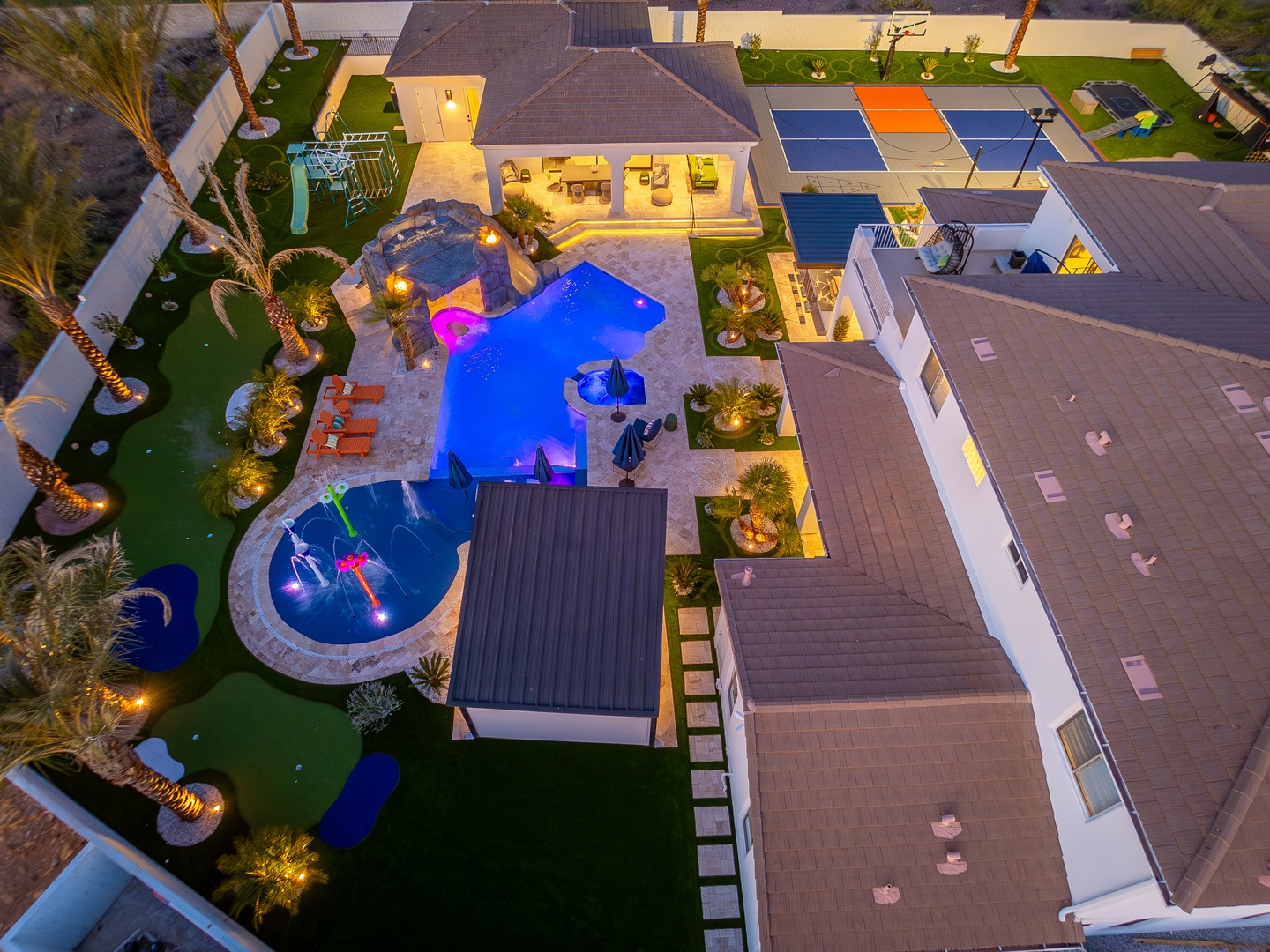 An expansive vacation property showcasing an illuminated freeform pool, vibrant splash pad, sports courts, and tropical landscaping—a private resort-style escape with exceptional outdoor living.