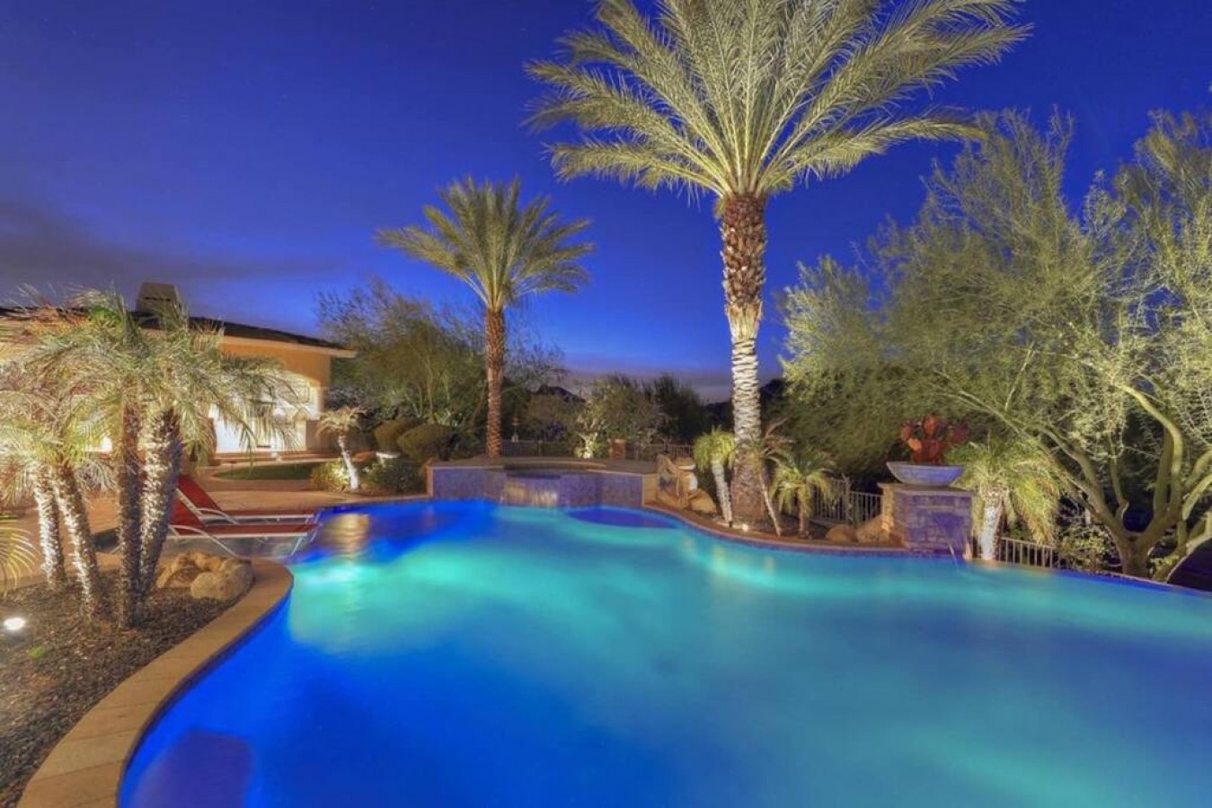 The property features a resort-style pool illuminated under a twilight sky, surrounded by mature palm trees and lush desert landscaping.
