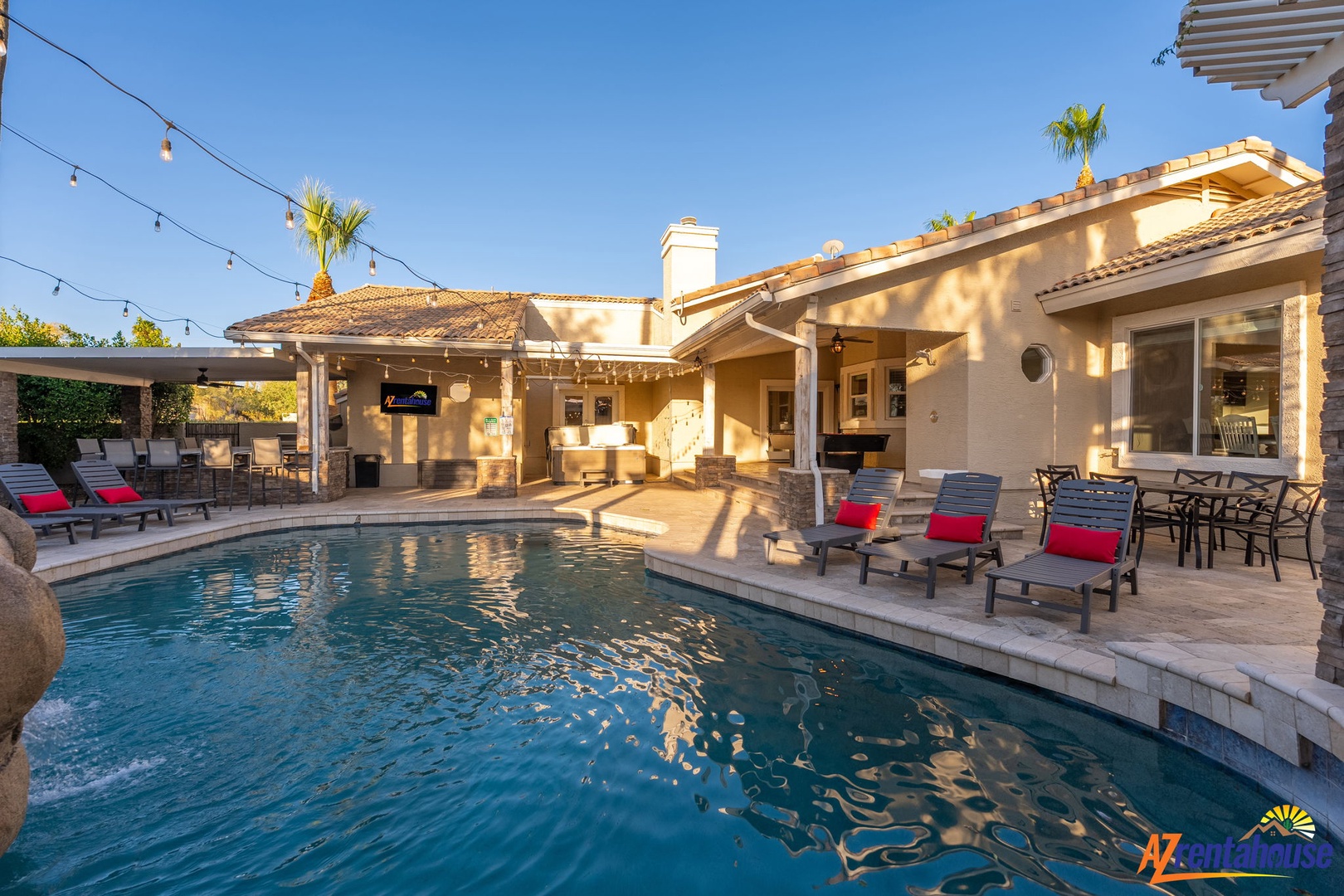 Stunning sun-drenched pool area framed by a beautifully designed desert oasis, featuring a covered outdoor lounge, dining space, and Spanish-tile architecture under string lights.