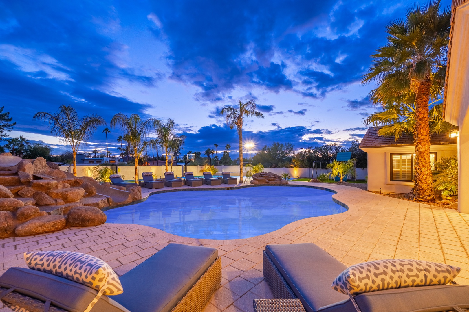 Stunning desert oasis featuring a resort-style pool with waterfall, framed by swaying palms and warm lighting beneath vibrant Arizona skies.