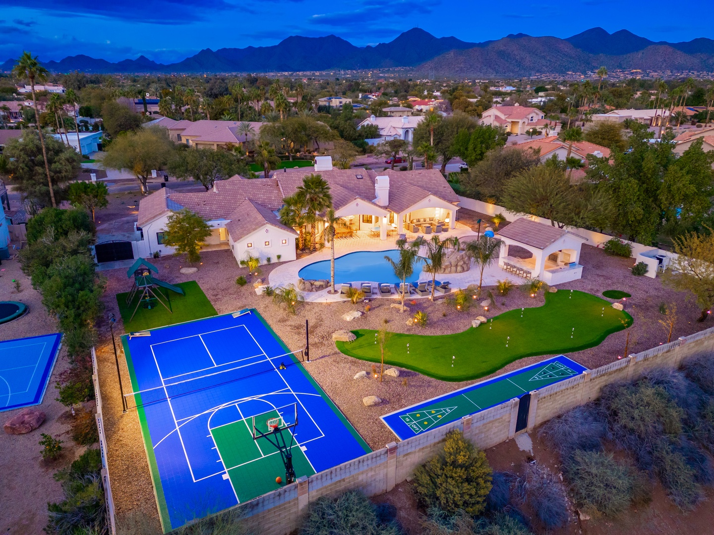 Aerial view of an expansive vacation estate featuring a sparkling pool, private tennis and sport courts, mini golf, and manicured desert landscaping, nestled in a residential neighborhood with mountain vistas.