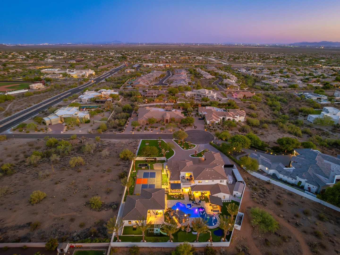 Aerial view of a spacious desert property with illuminated pool and patio, surrounded by native landscaping in a quiet residential neighborhood at dusk.