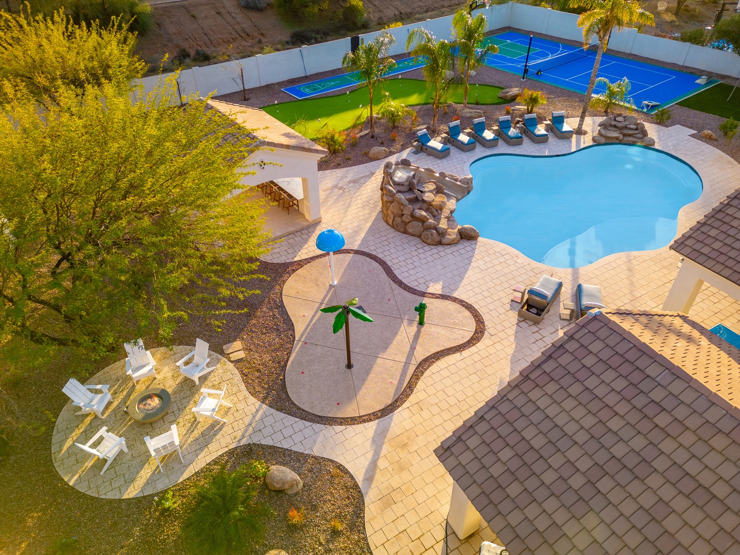 Aerial view showcasing the property's extensive recreational amenities, including a resort-style pool with waterfall feature, splash pad, tennis court, lush lawns, and shaded outdoor seating areas surrounded by desert landscaping.