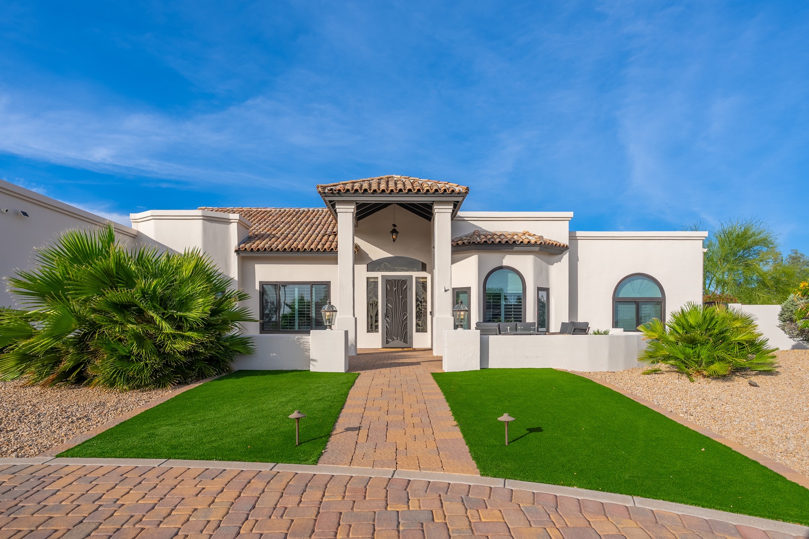 Elegant Mediterranean-style villa with terra-cotta tile roof and arched entrance, nestled in a lush desert landscape with vibrant blue skies overhead.